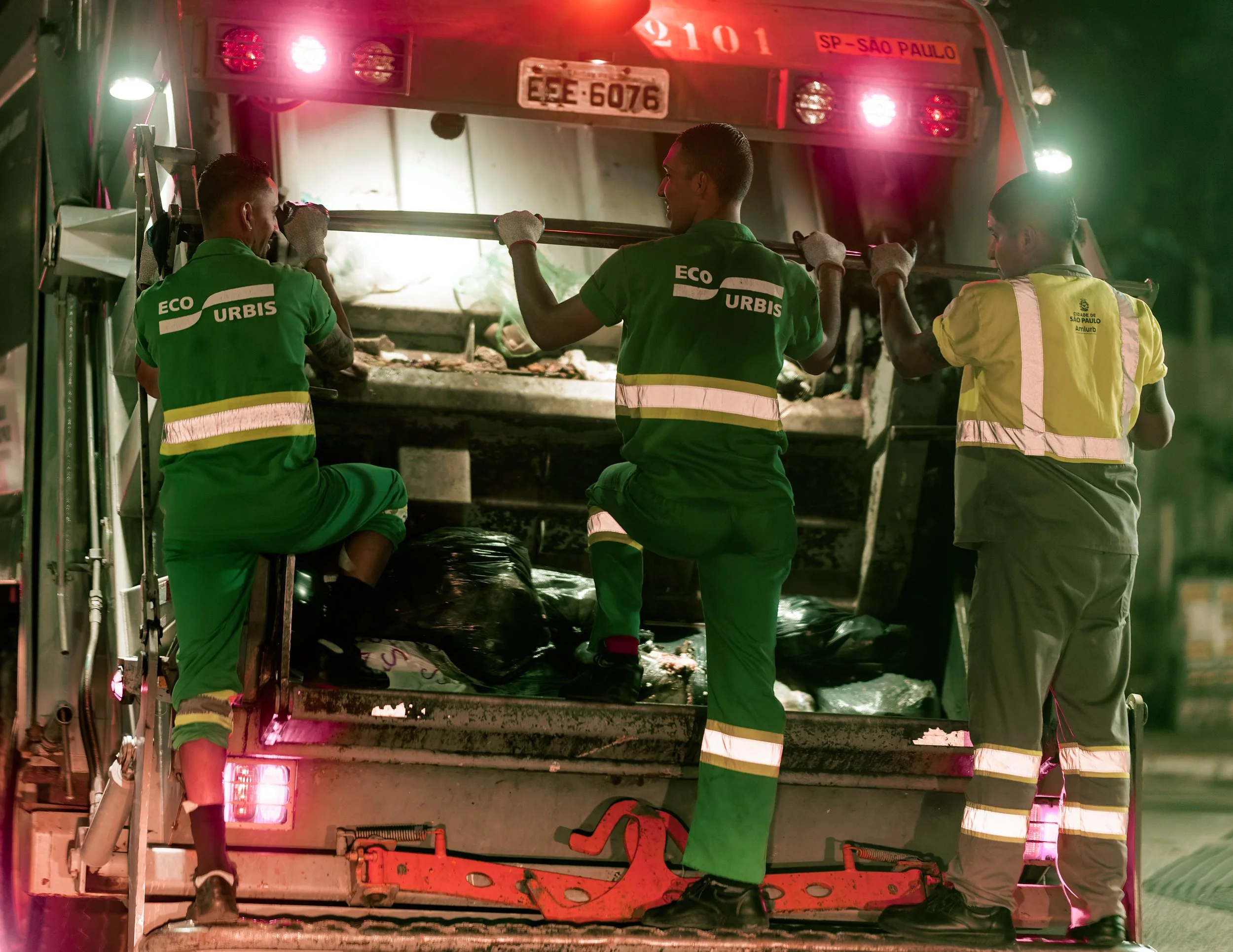 Three firefighters in green and yellow uniforms working together to open the back of a fire truck at night. The back of the truck has a license plate 'ESE 6076' and a label 'SP-SAO PAULO', with red and white lights illuminating the scene.