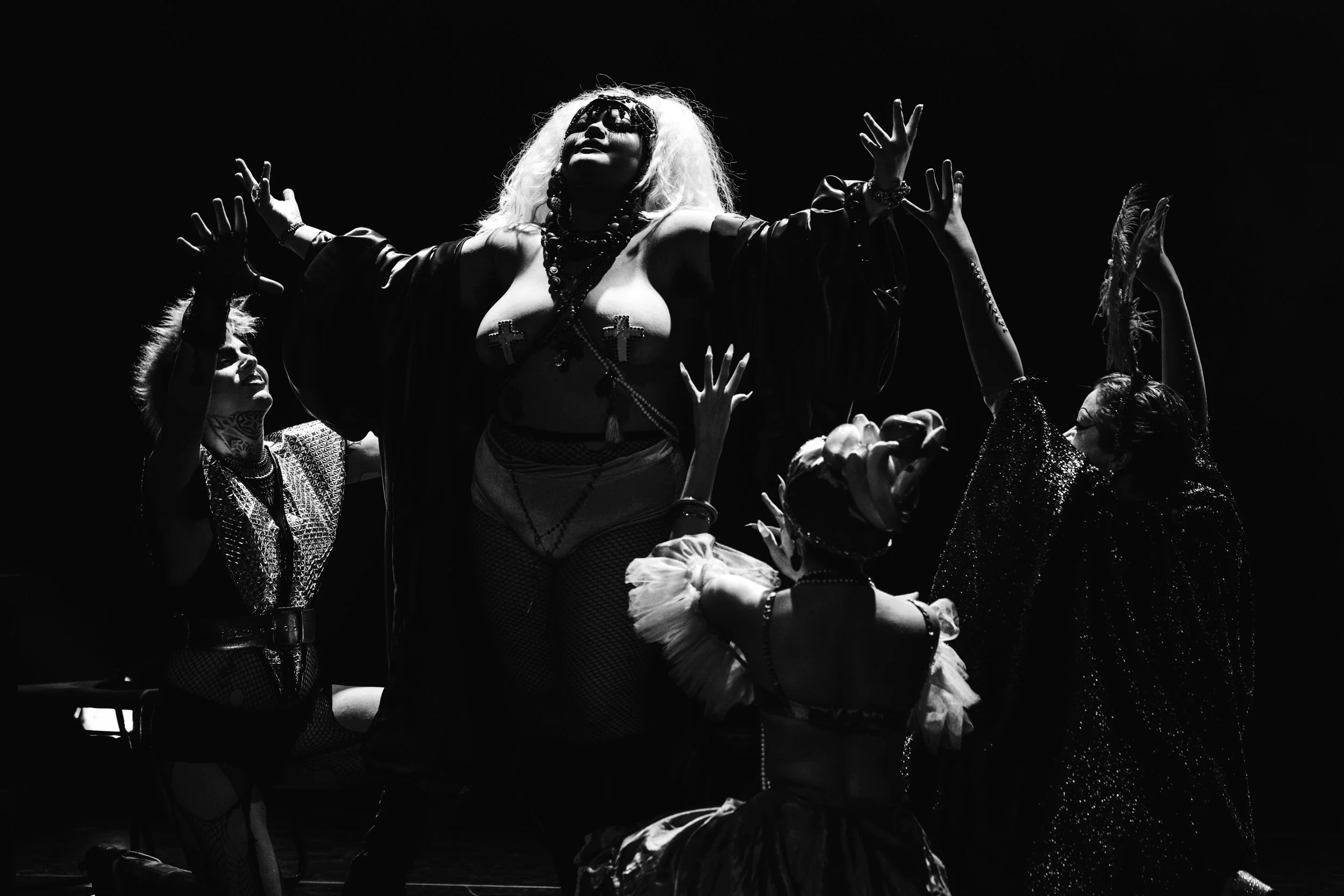 A black and white photo of a performer with long, curly hair and dark sunglasses, wearing a revealing outfit decorated with crosses, on stage with several hands reaching up towards her, suggesting she is the center of attention.
