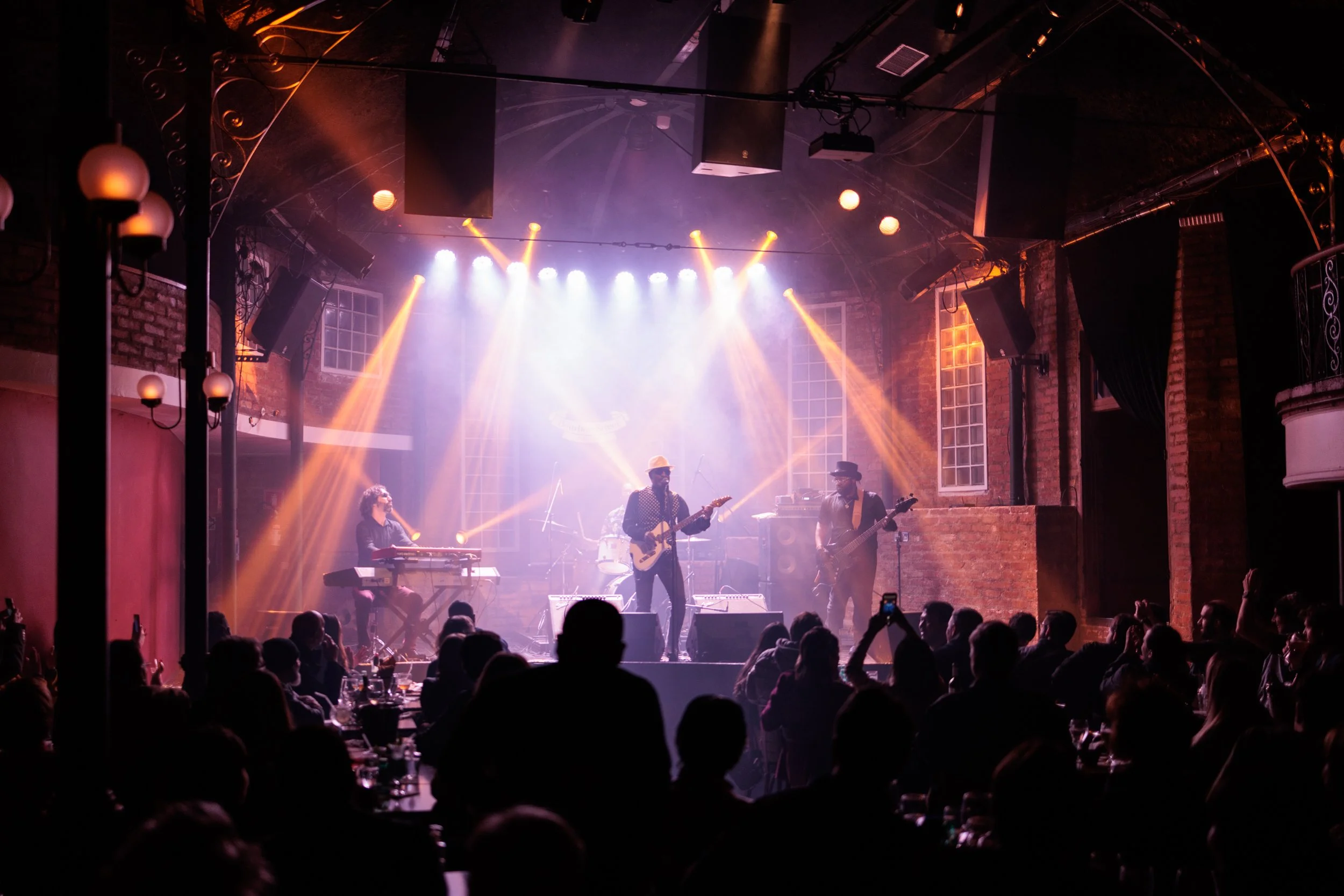 A live music performance on stage in a dimly lit venue with colorful stage lights, including a keyboardist, two guitarists, and an audience watching and taking photos.