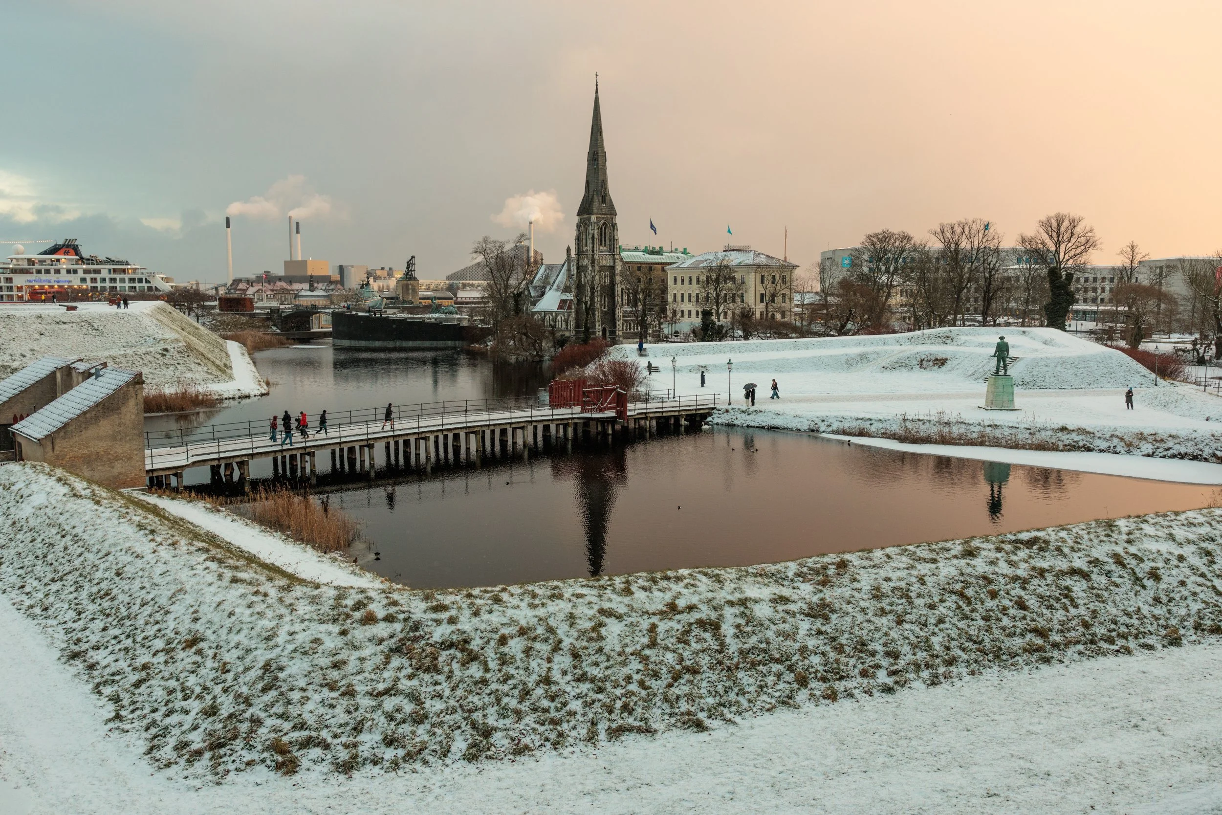 A snowy waterfront park with a bridge, walking path, and statue, alongside buildings, a church with a tall spire, and a cruise ship in the background.
