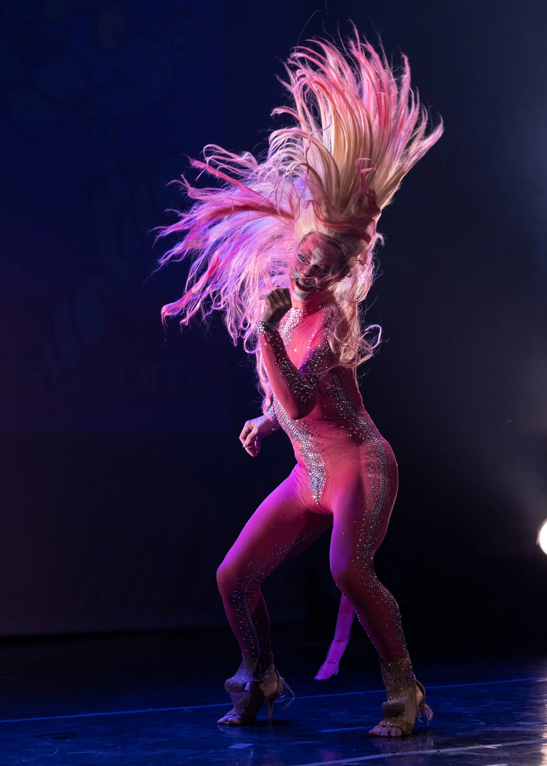 A performer in a vibrant pink costume with a large, feathered headdress dances on stage under dark lighting.