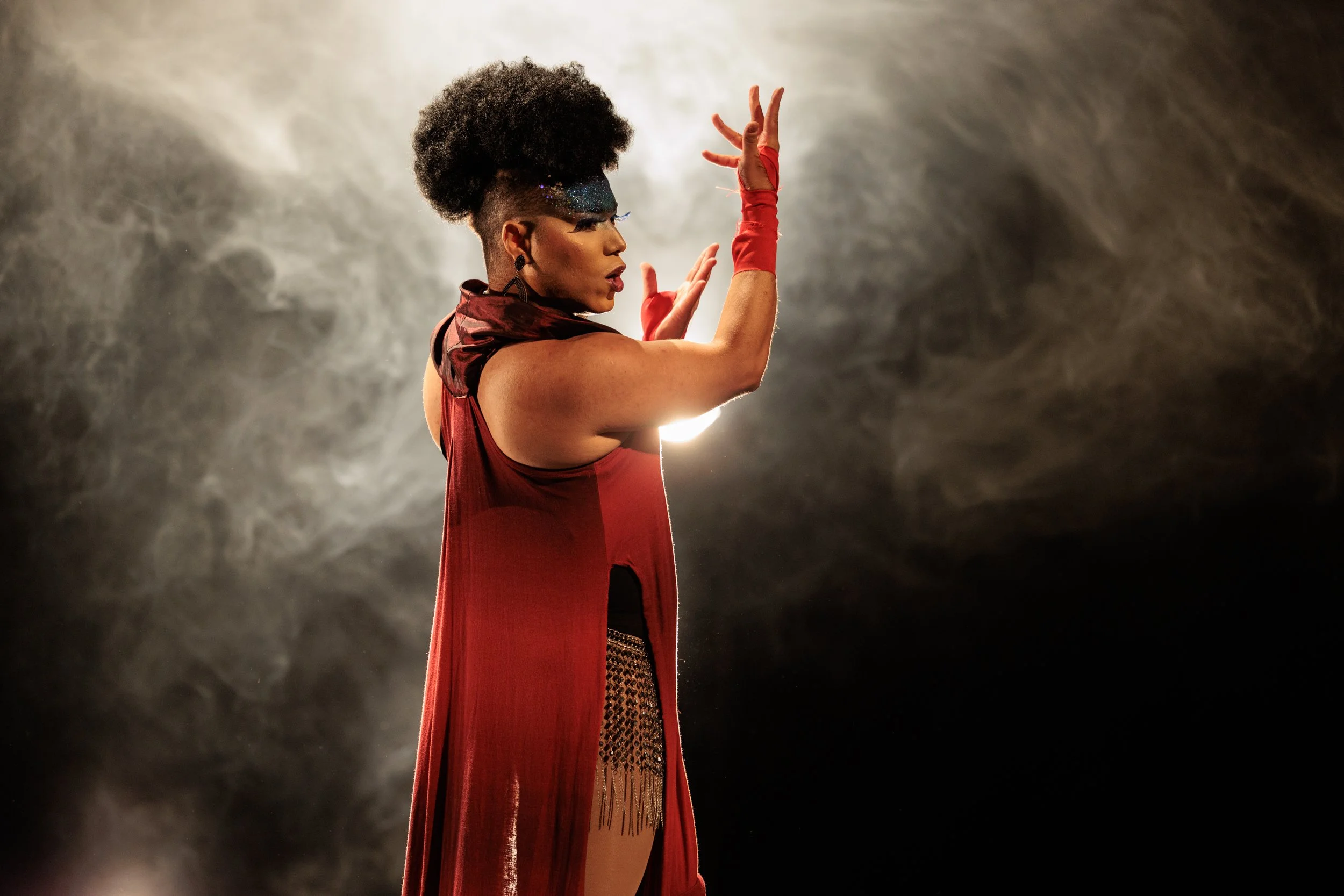 A performer with styled hair, vibrant makeup, and a red outfit poses against a smoky background with a dramatic light behind her.