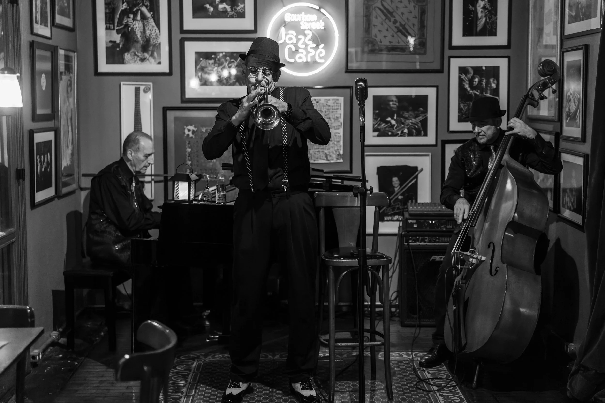 Black and white photo of a jazz band performing in a cozy cafe. The band consists of a notable looking trumpet player, a pianist, and a double bass player. The background has framed pictures and a neon sign that says 'Jazz Cafe.' The scene is intimat