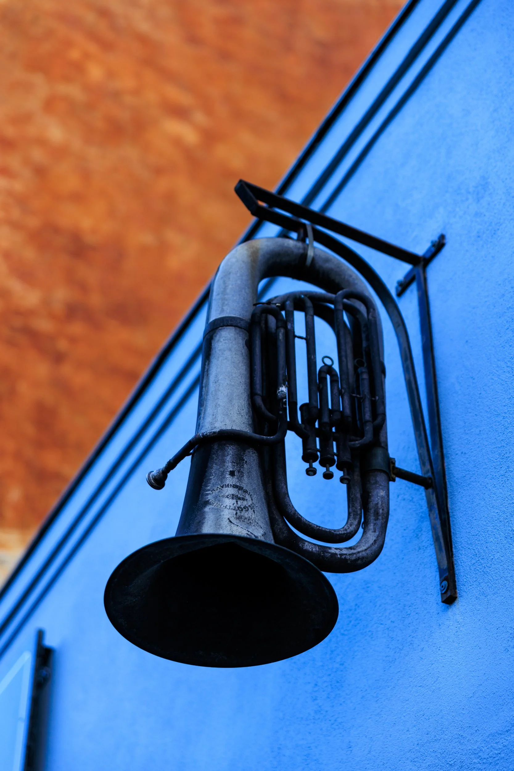 Close-up of a brass industrial horn mounted on a blue wall, with a curved pipe and black brackets, and a blurred wooden surface in the background.