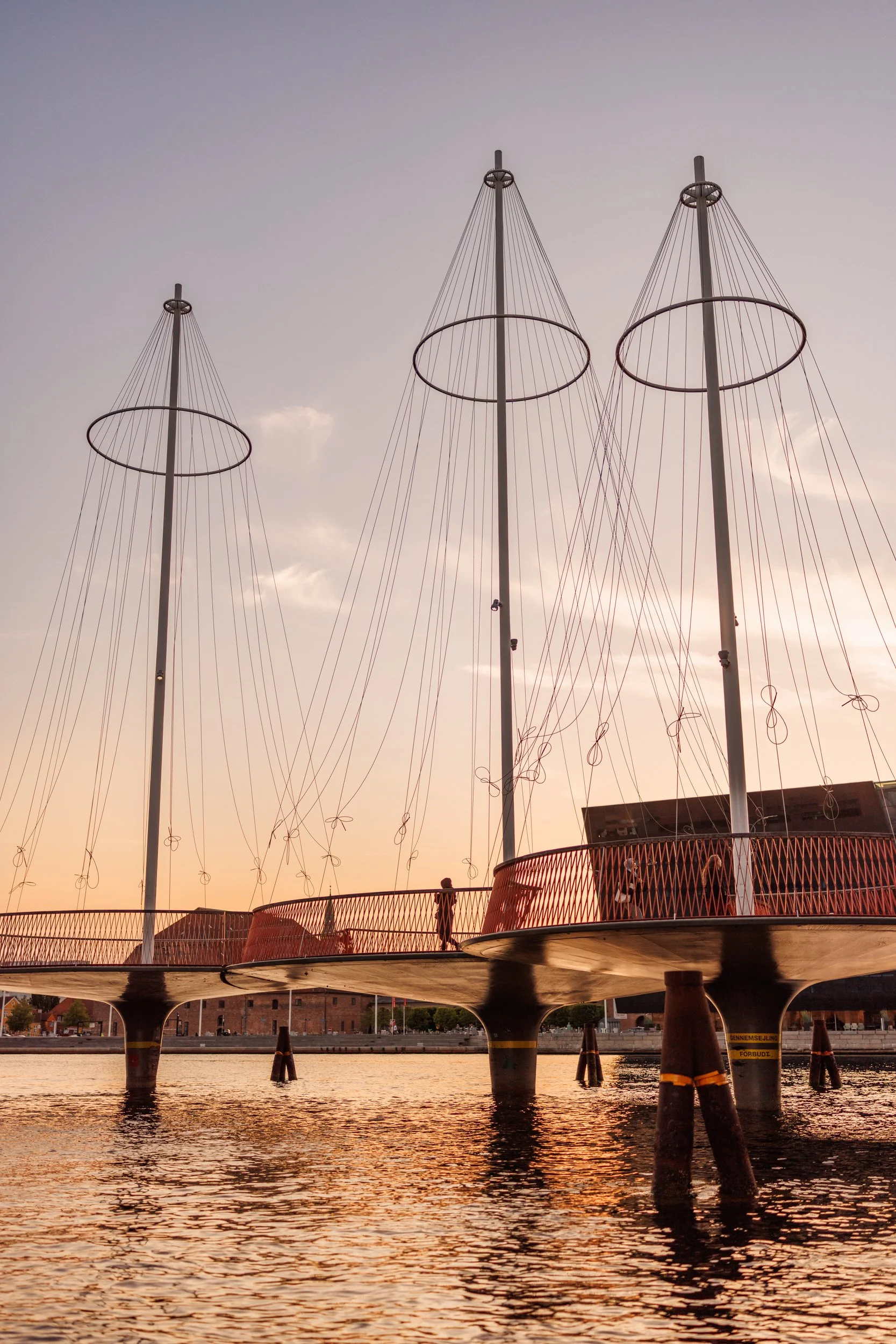 Tall poles with circular structures and string-like elements hang down, forming the shape of a sailboat mast and rigging. The scene is set during sunset over a body of water with reflections of the sky and a small construction platform or tower under