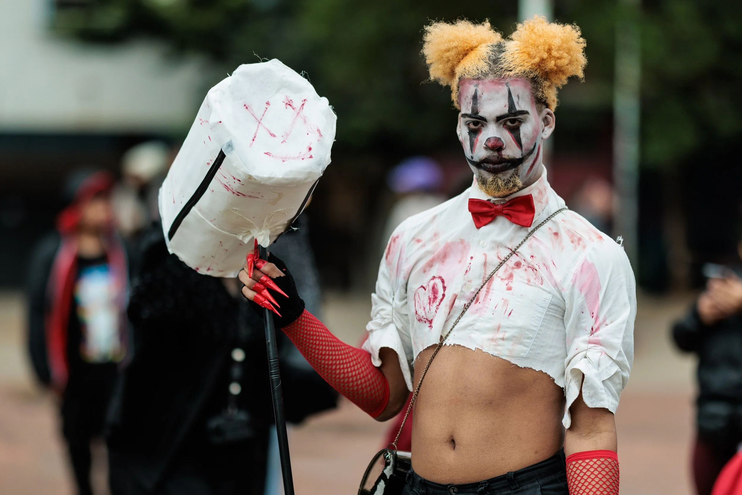 Person dressed as a creepy clown with painted face, curly blond hair in pigtails, and a red bow tie, holding a white balloon with a smiley face drawn on it, at an outdoor event or parade.