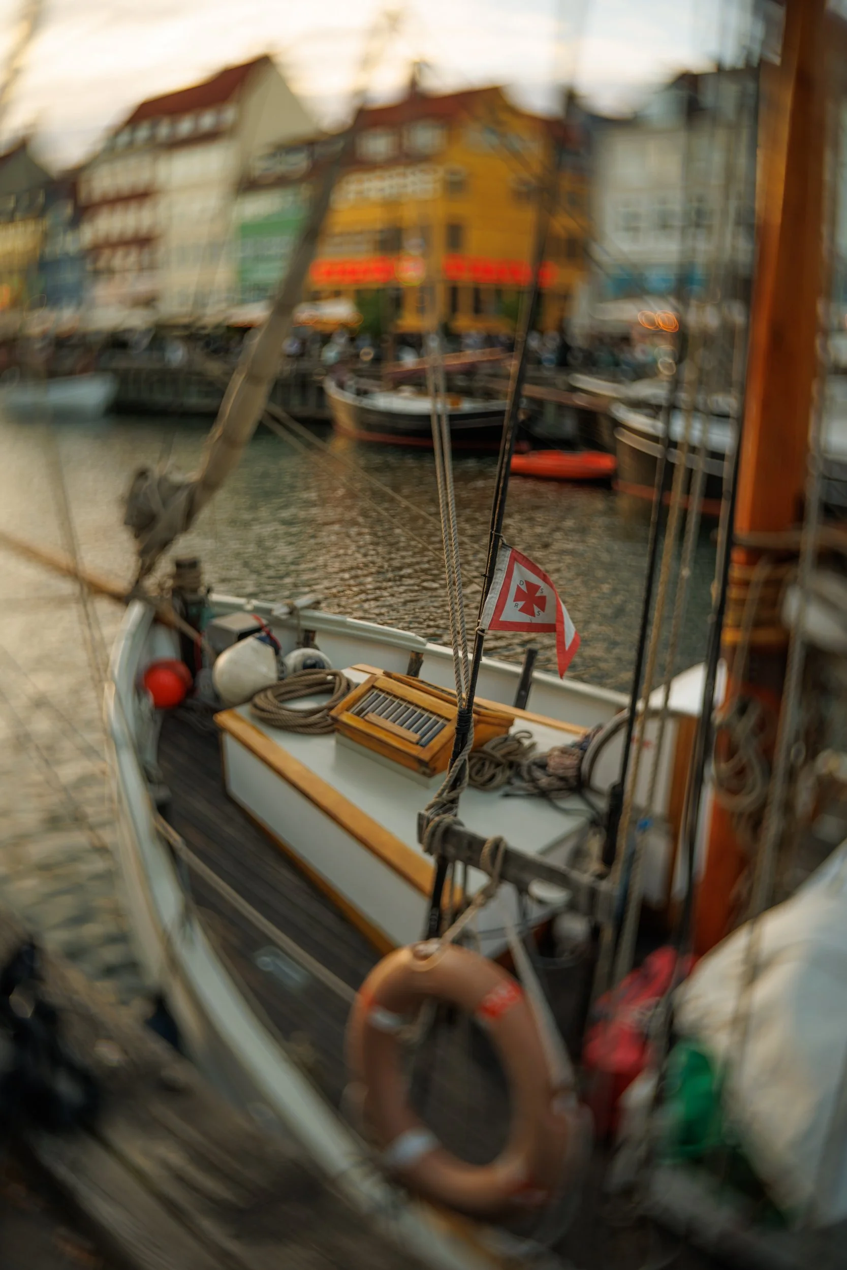 Blurry photo of a boat's interior with ropes, a toolbox, and a small red and white flag, with a view of colorful buildings and boats along a waterfront.
