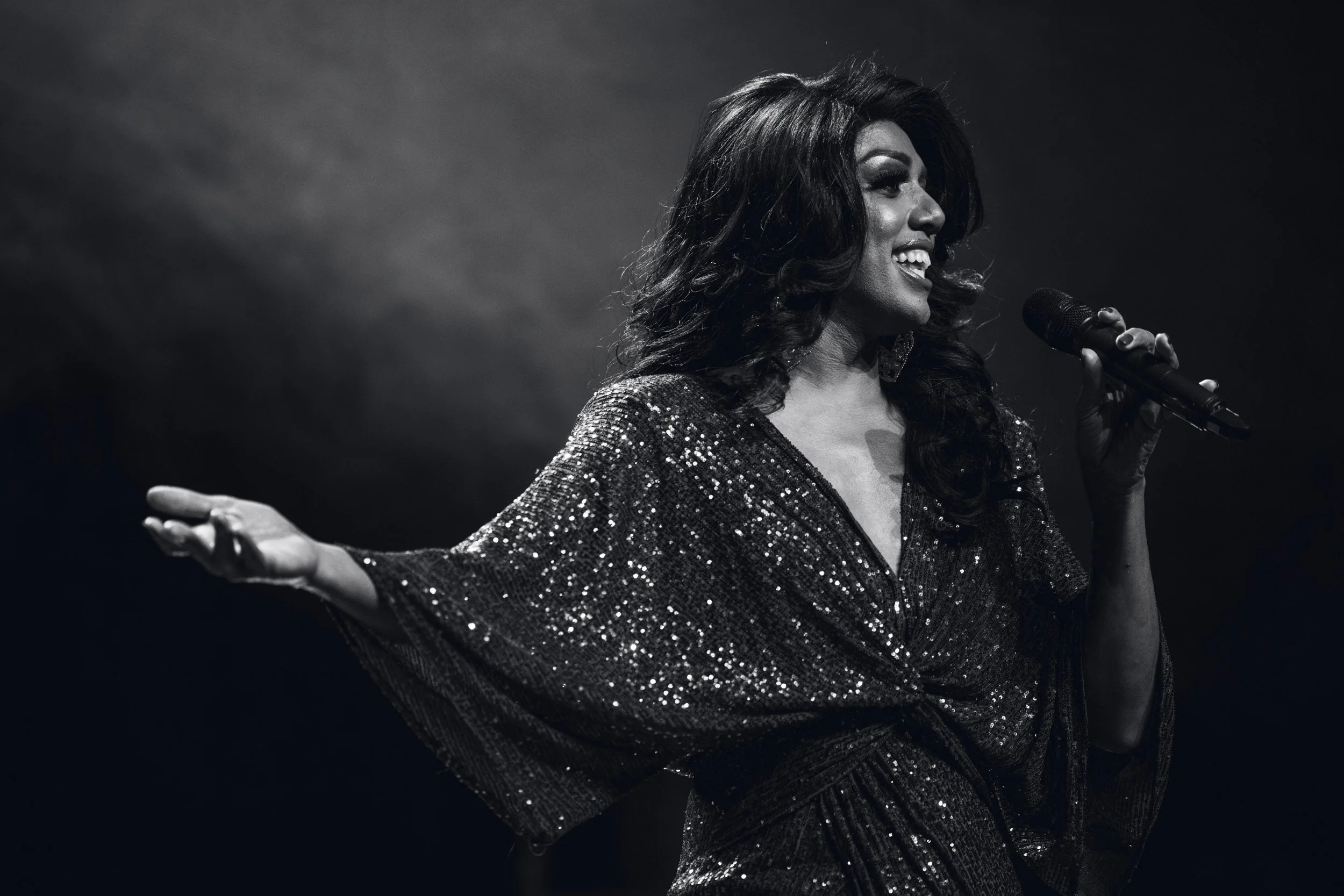 A woman singing into a microphone with her arms outstretched, wearing a sparkling dress, smiling, in a black and white photo.