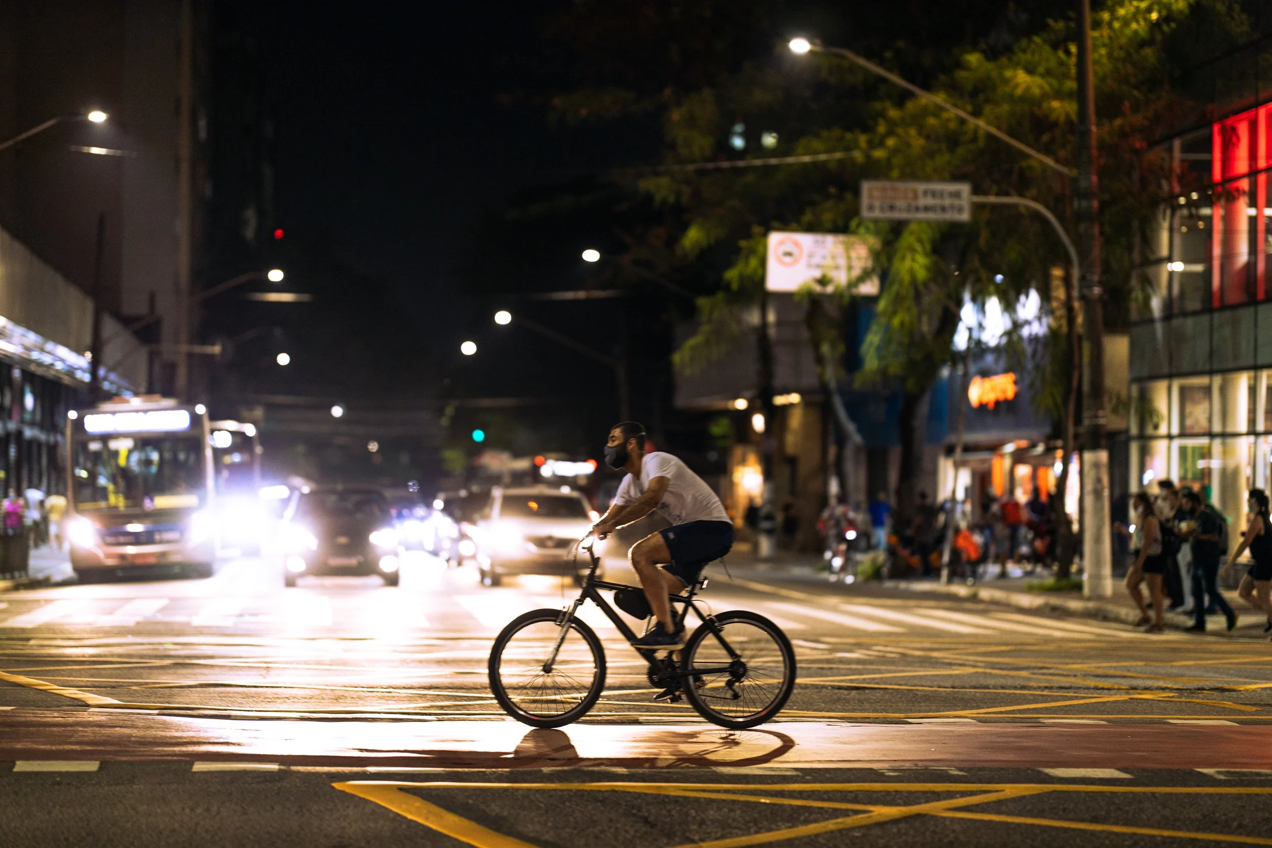 A person wearing a face mask riding a bicycle across a city street at night, with cars and illuminated buildings in the background.