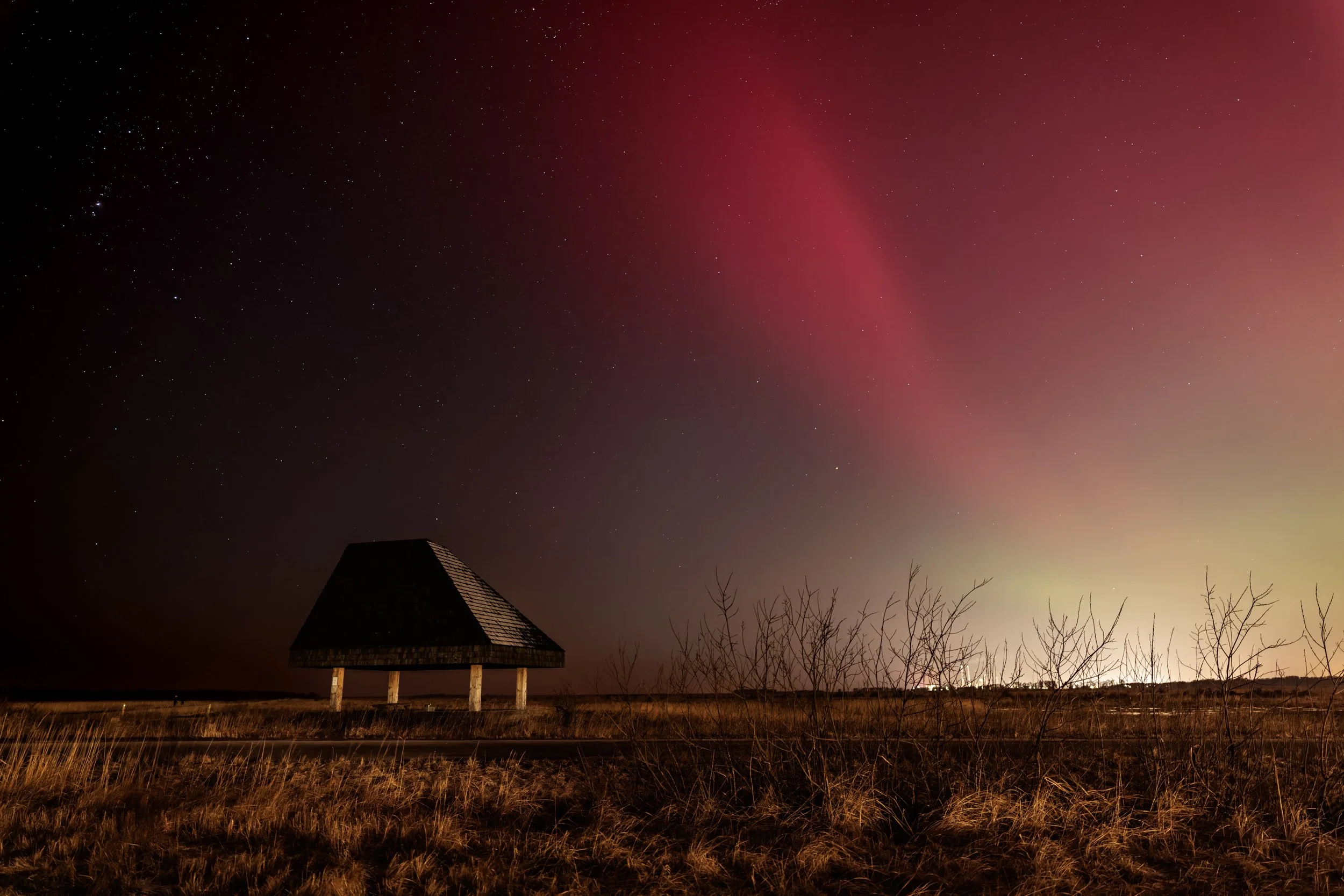 Night sky with countless stars and a reddish aurora arching across the horizon. A small, dark, roofed structure on stilts is visible in the foreground, along with some leafless trees and dry grass.