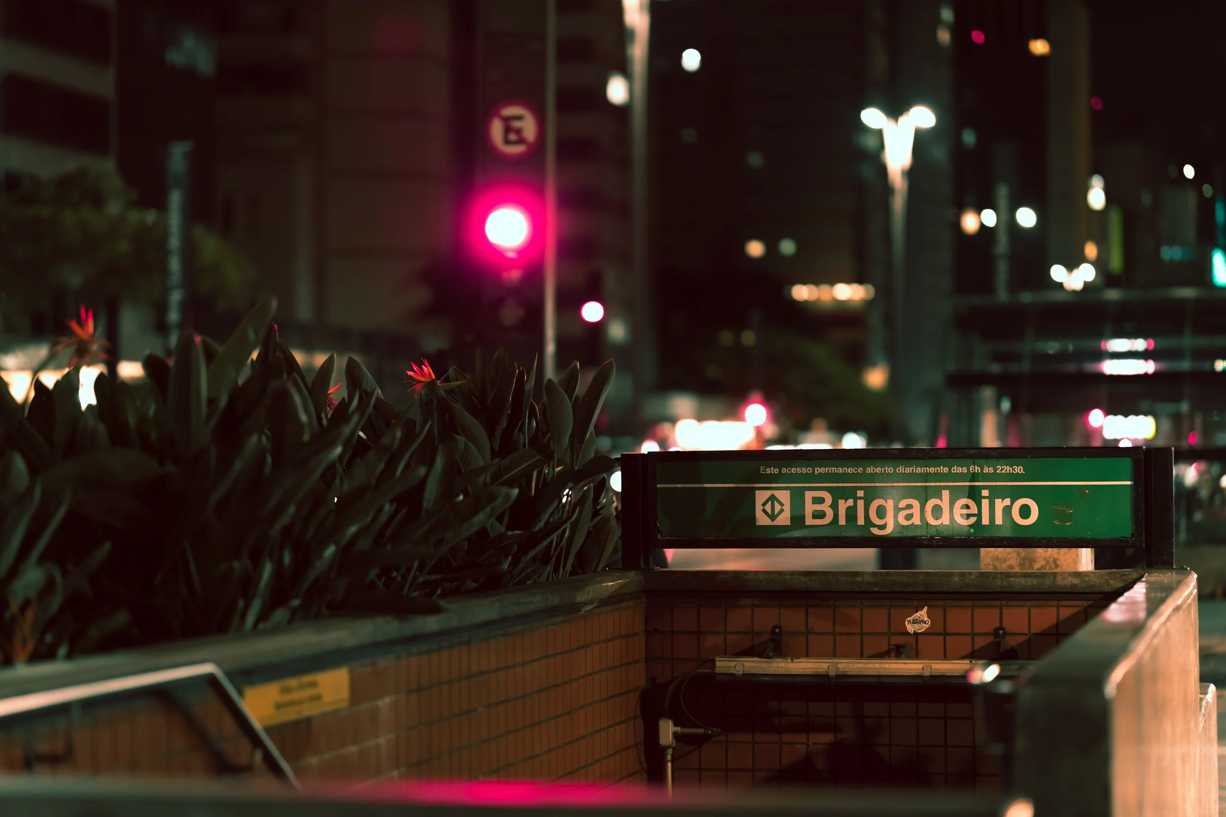 Night scene at a city subway station with green sign reading "Brigadeiro" in Portuguese, lit pink traffic signal, in the background blurred city lights and buildings, and plants in the foreground.