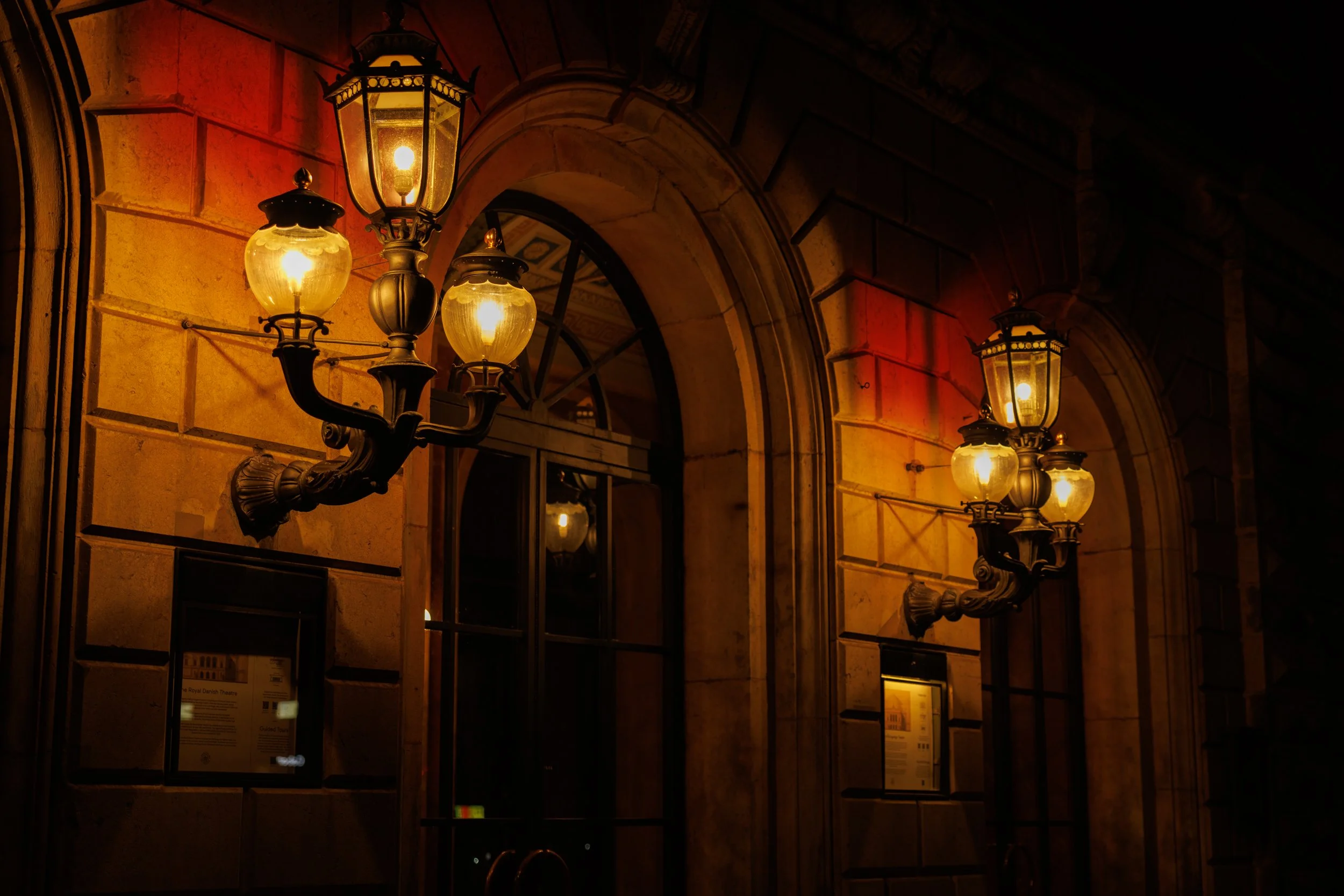 A row of ornate street lamps attached to a stone building exterior at night, illuminating the area with warm light.