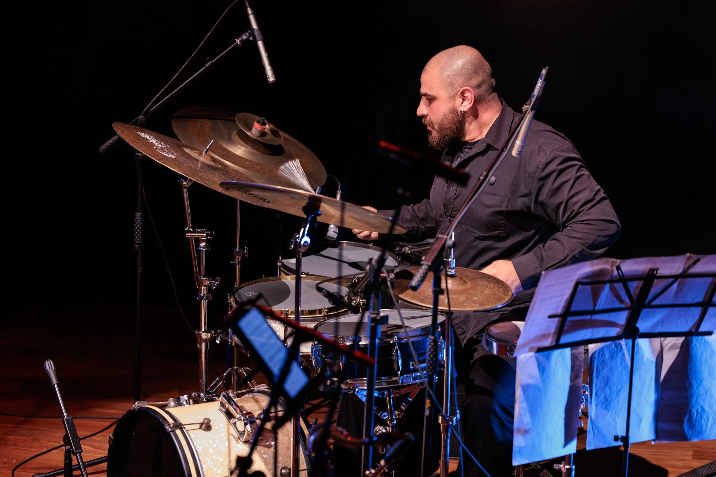 A man playing drums on stage with black background, wearing a dark shirt, and a music stand with sheet music in front of him.