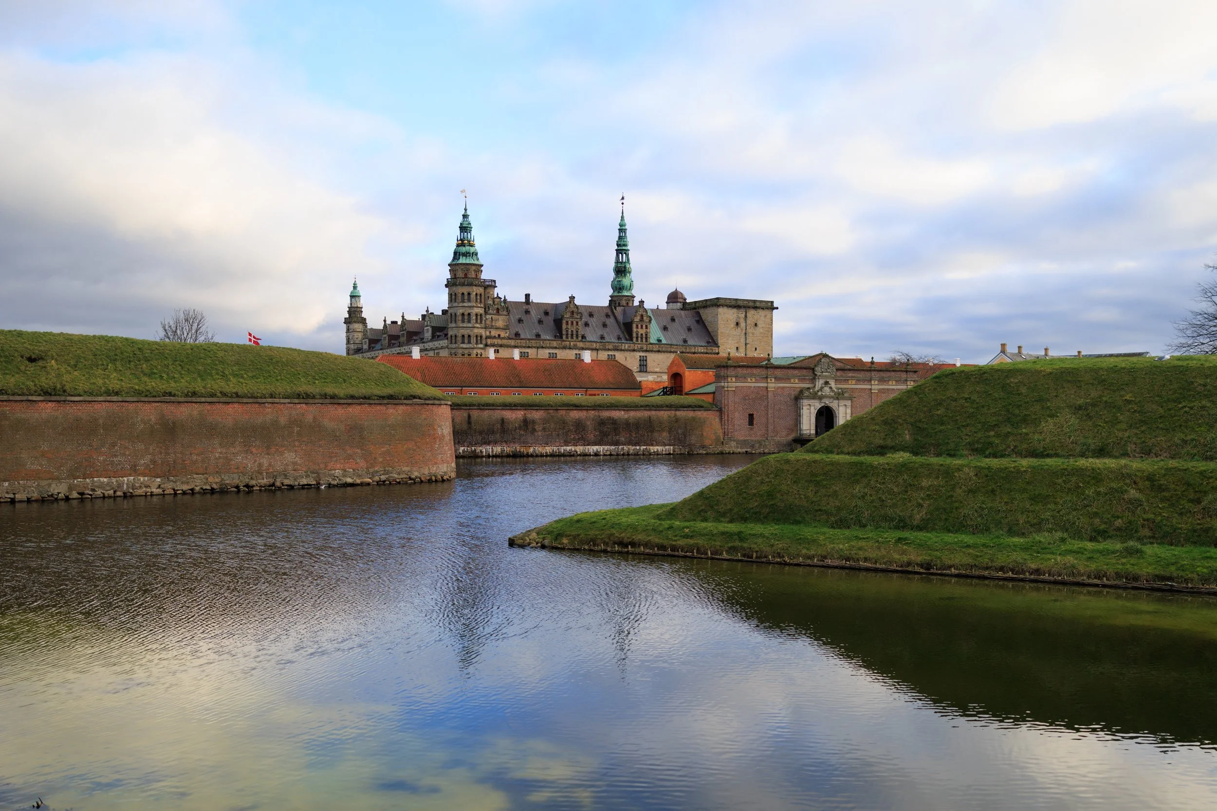 View of a historic castle with towers and spires across a body of water, with grassy banks and a cloudy sky.
