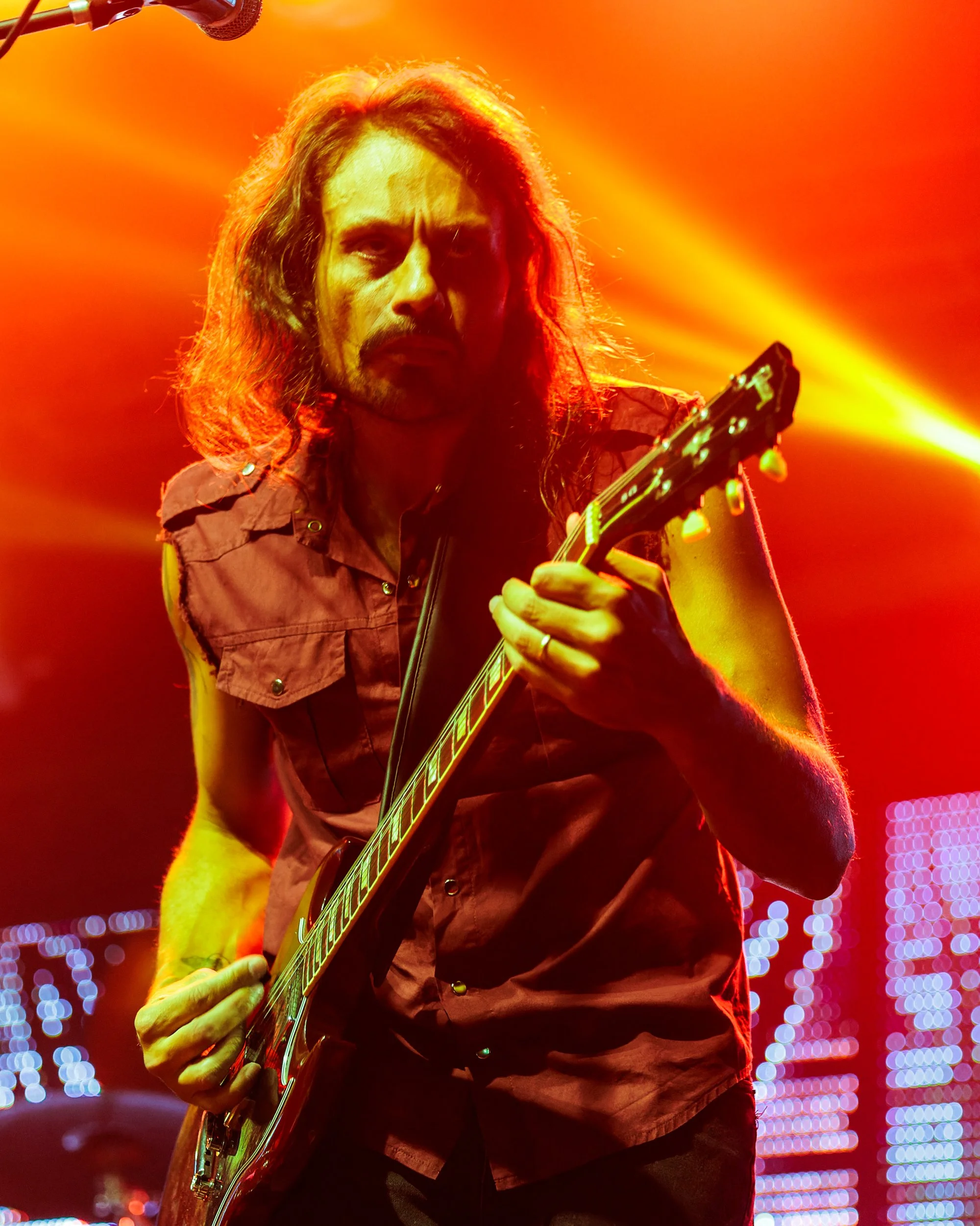 A male musician playing an electric guitar on stage, illuminated by red and orange stage lights, with a blurred background of colorful lights.