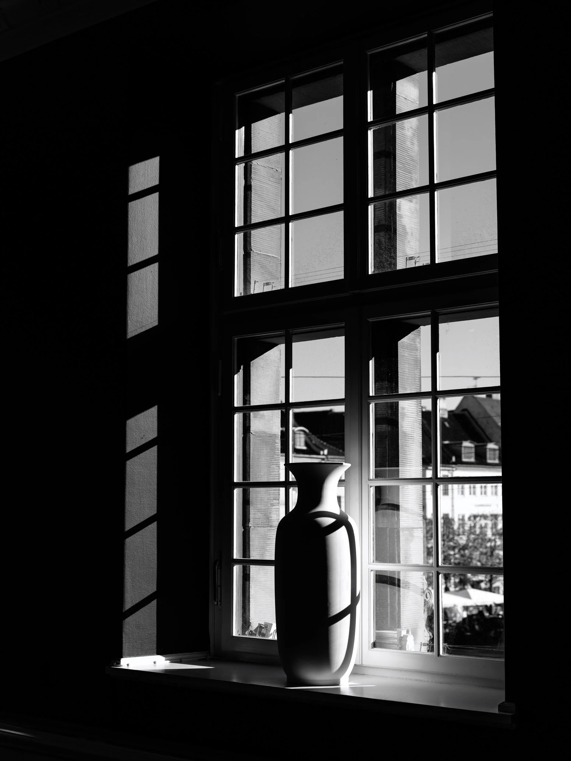 A black and white photograph of a view through a window, with sunlight casting shadows of the window frame onto the interior wall. A decorative vase sits on the windowsill, and buildings are visible outside.
