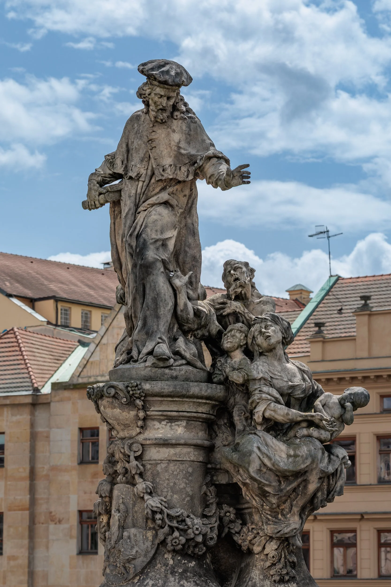 A stone sculpture featuring several figures, with a prominent central figure wearing a cap and holding a staff. Surrounding figures appear to be engaged in expressive poses, set against a backdrop of rooftops and a partly cloudy sky.