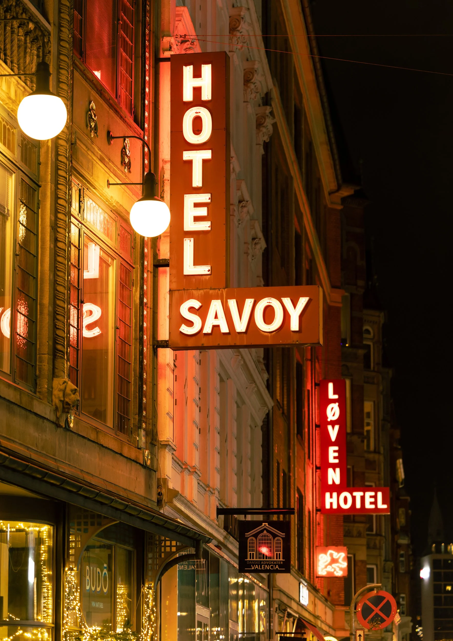Nighttime view of city street with illuminated signs for a hotel and various businesses. Neon signs including 'HOTEL', 'SAVOY', and others are visible, along with street lamps and building facades decorated with lights.