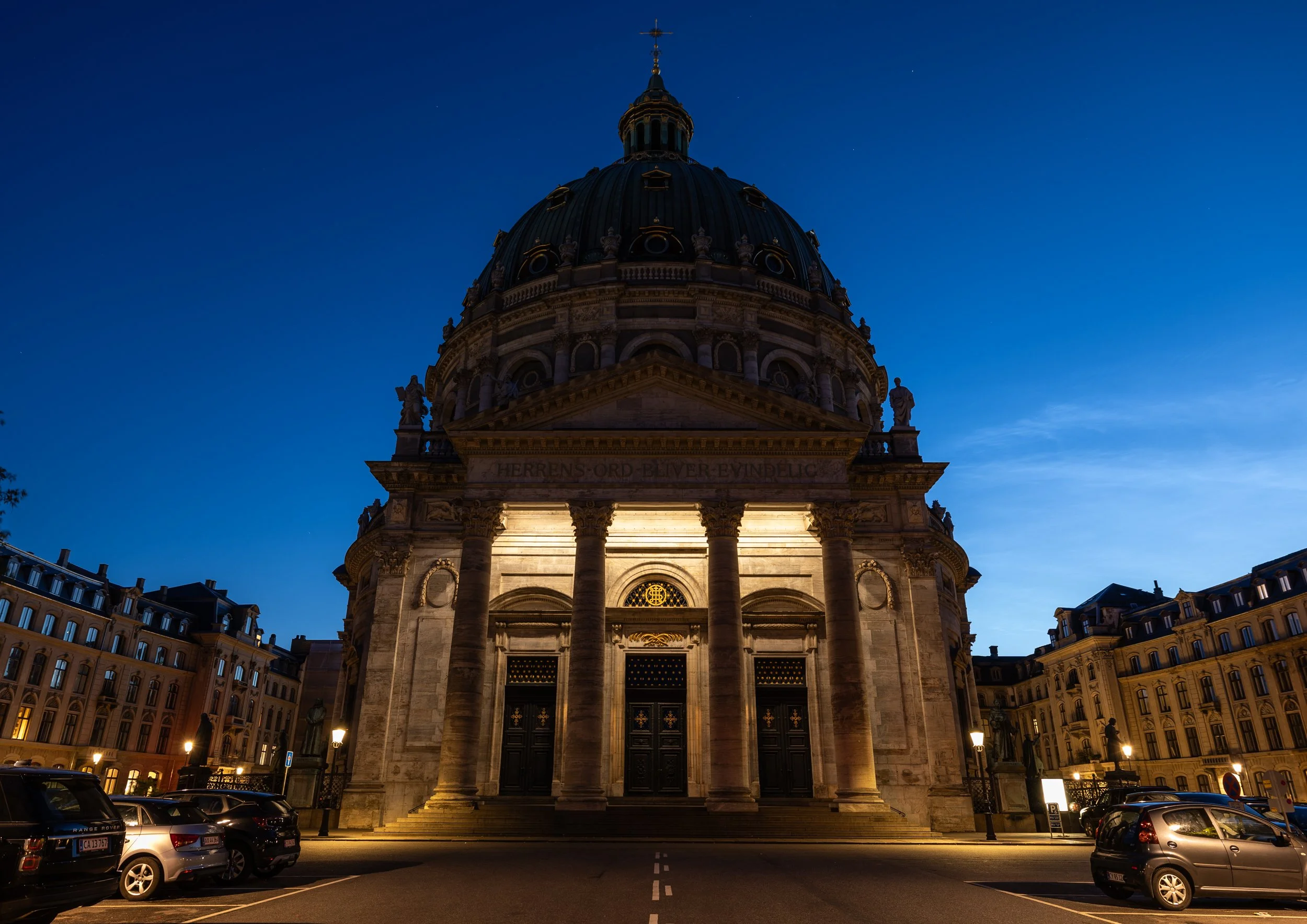 A historic building with a large dome illuminated at night, streets filled with parked cars, and surrounding classical architecture, under a clear dark blue sky with stars.