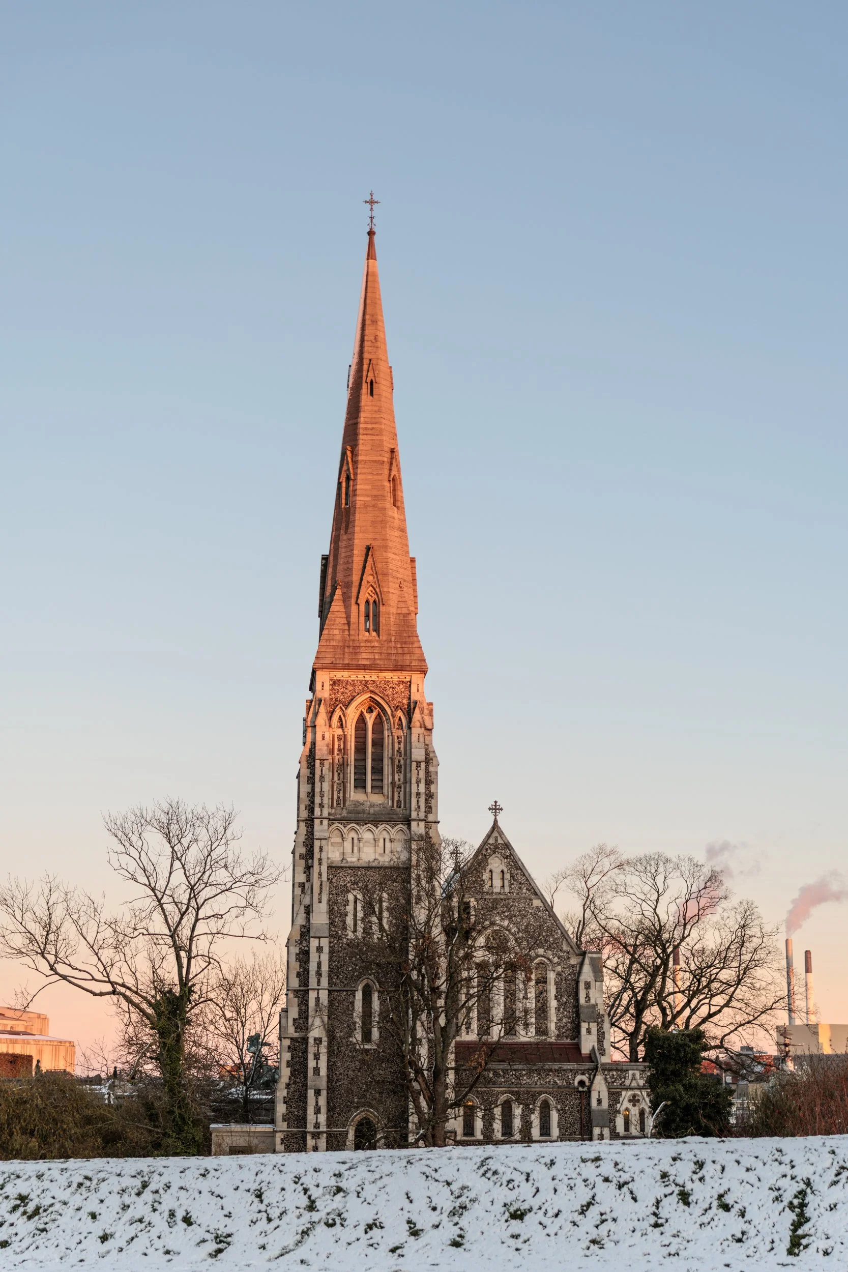 A tall church steeple with a cross at the top, set against a clear evening sky. The church has stained glass windows and stone architecture, with leafless trees in the foreground and snow on the ground.