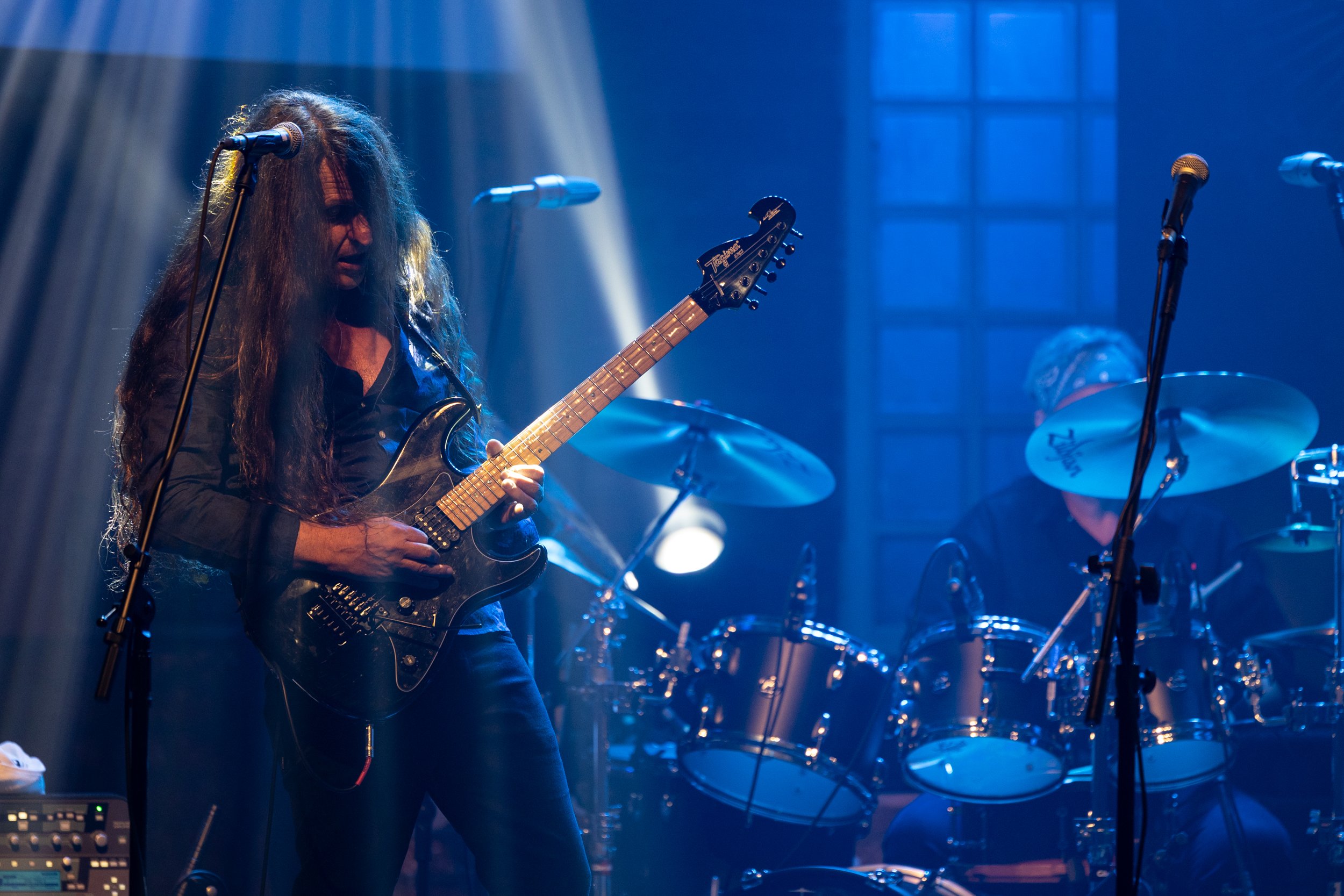 Musician playing an electric guitar on stage with drums in the background, stage lights creating a dramatic atmosphere.