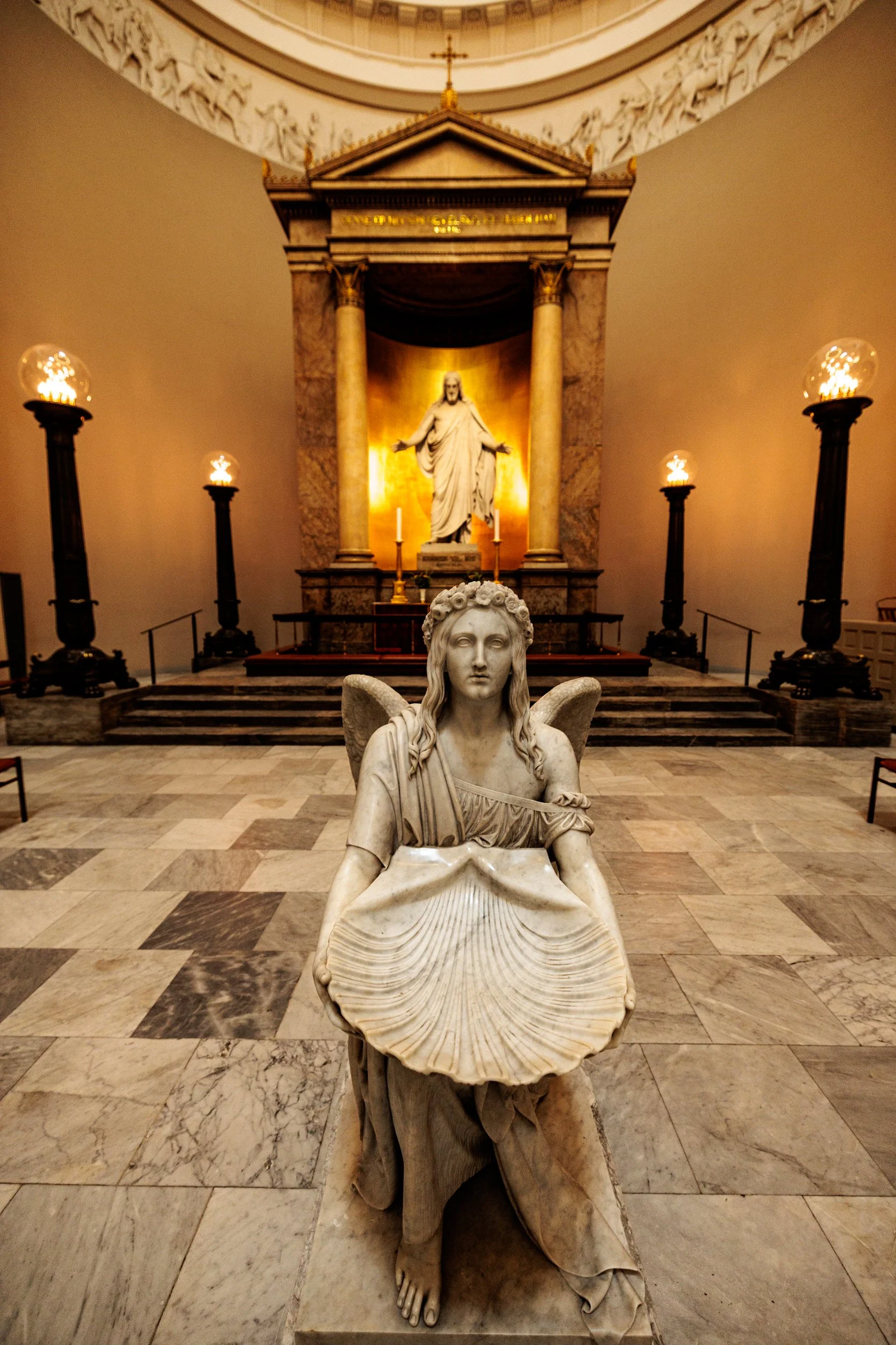 Marble statue of a woman with a shell in her lap, seated with one leg crossed over the other, in a church or cathedral interior, with a background altar featuring a statue of Jesus Christ, candles, columns, and ornate architectural details.
