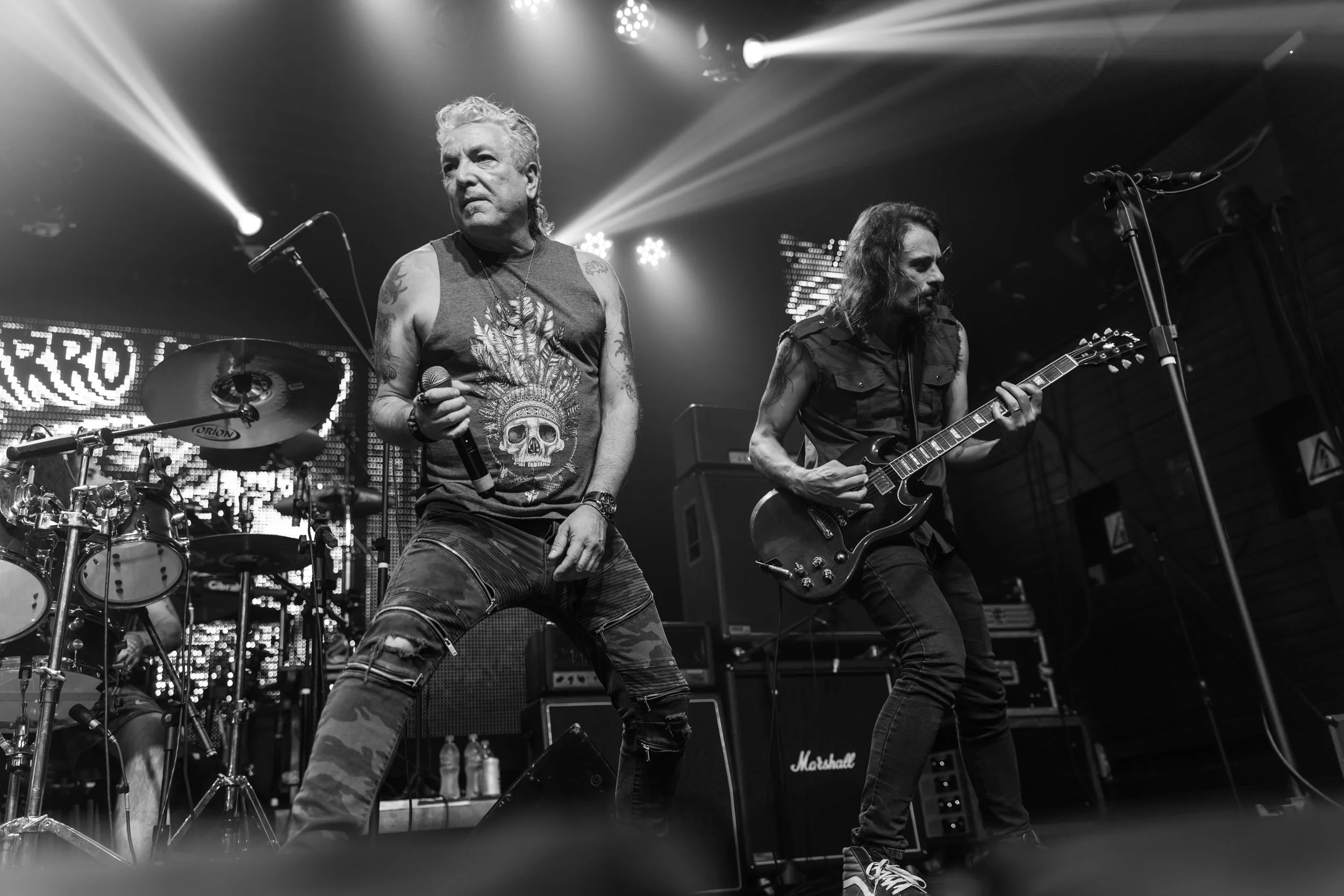 Black and white photo of two male musicians performing on stage. One is singing into a microphone and the other is playing an electric guitar. The stage is lit with bright spotlights and there is a drum set in the background.