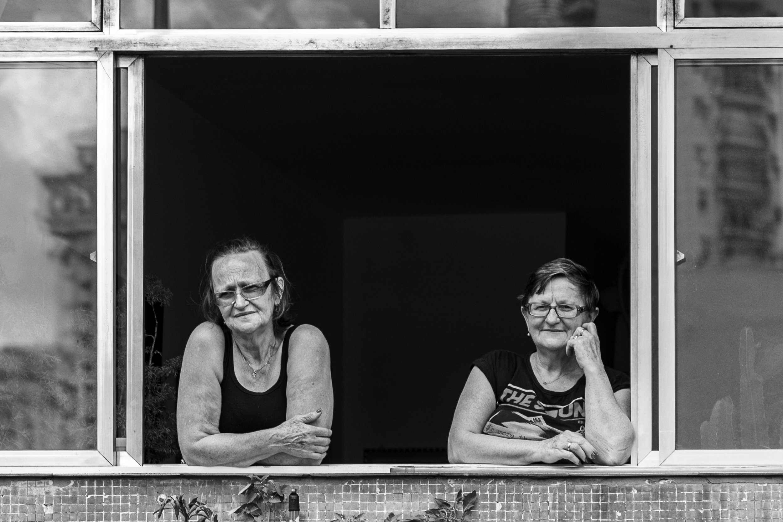 Two women leaning on a windowsill, looking outside. The woman on the left has glasses, her hair tied back, and is wearing a tank top. The woman on the right also wears glasses, has short hair, and is wearing a graphic t-shirt. The window frame is metal, and the background is dark.