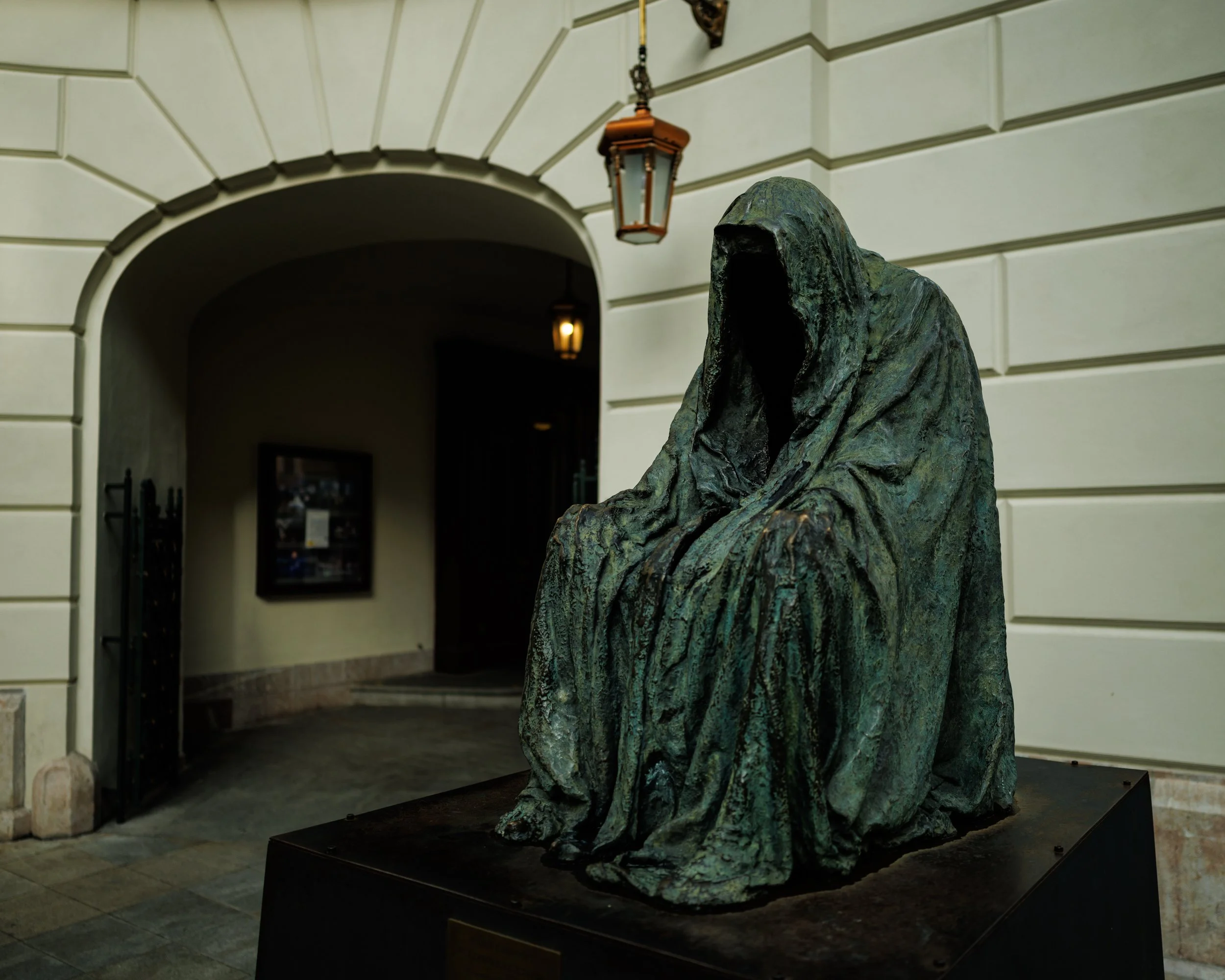 Bronze statue of a hooded, cloaked figure with no visible face, seated with hands resting on knees, placed indoors in front of a building with arched entryway and beige walls.