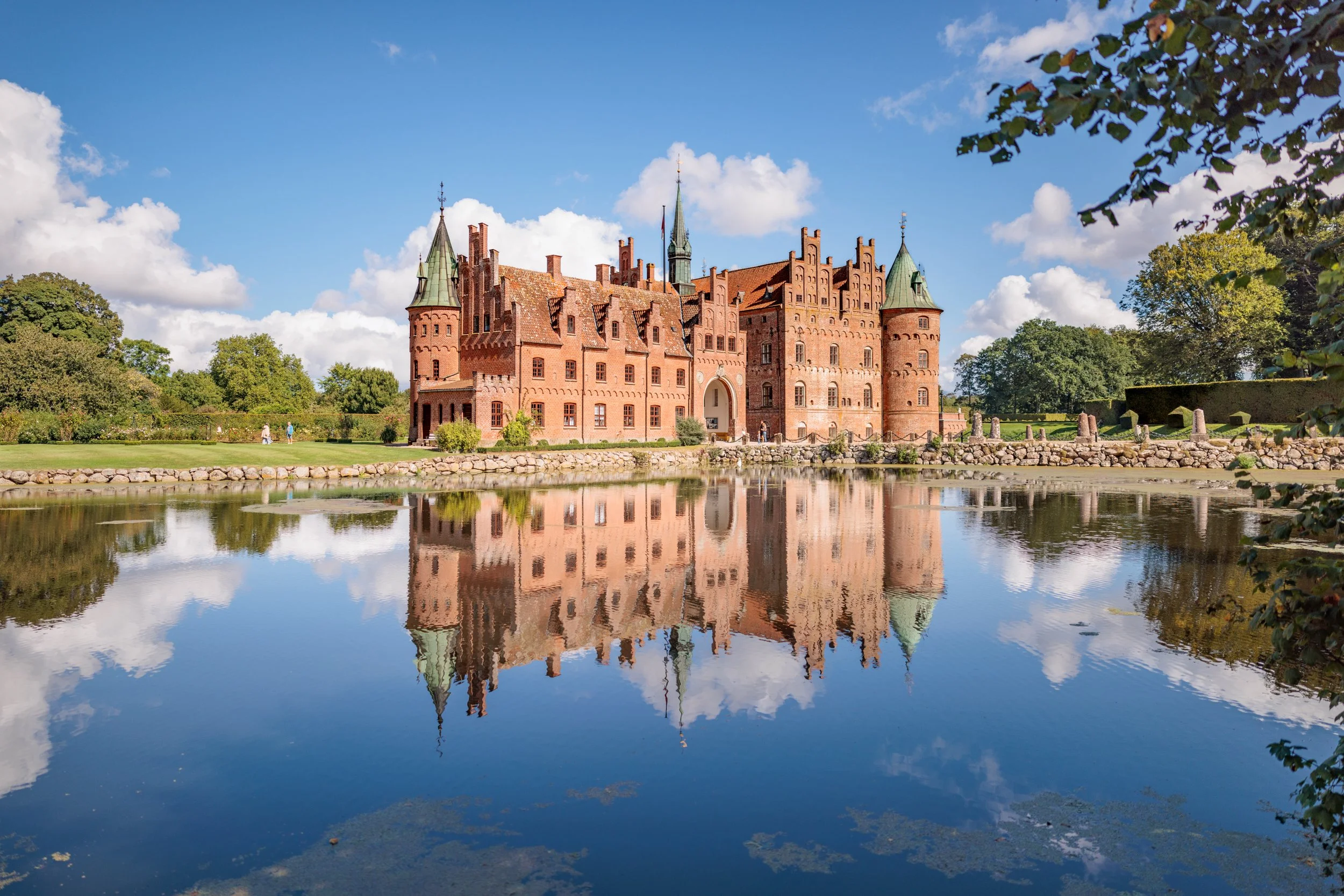 A historic red brick castle with green spires, surrounded by water reflecting the castle and a partly cloudy blue sky.