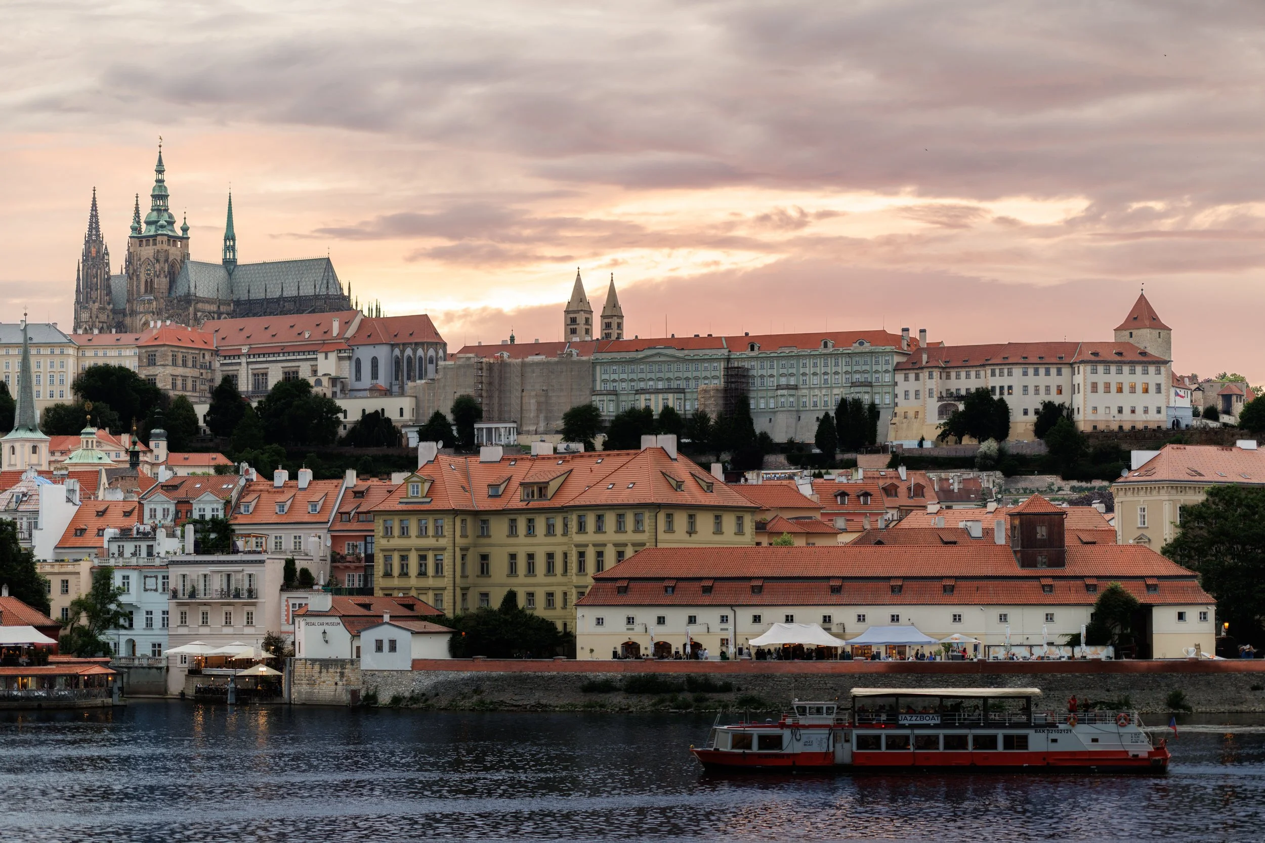 A cityscape with historic buildings and churches viewed from a body of water at sunset, with a boat labeled 'Jazzboat' in the foreground.