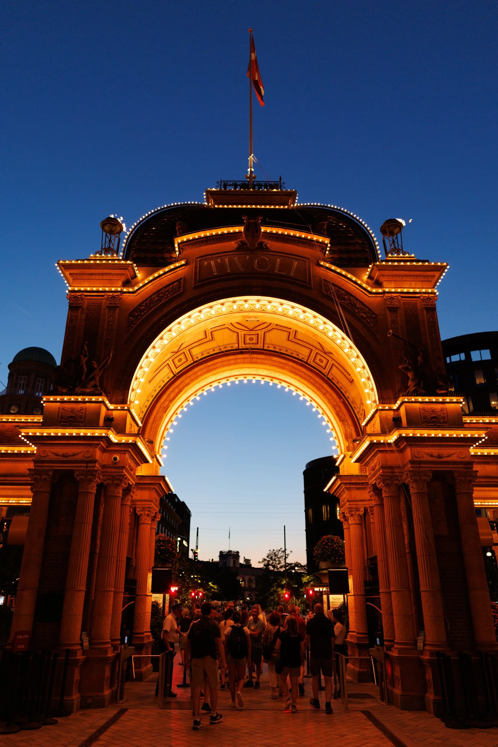 The illuminated archway at Tivoli Gardens in Copenhagen, Denmark, during early evening with people walking underneath and a clear sky in the background.