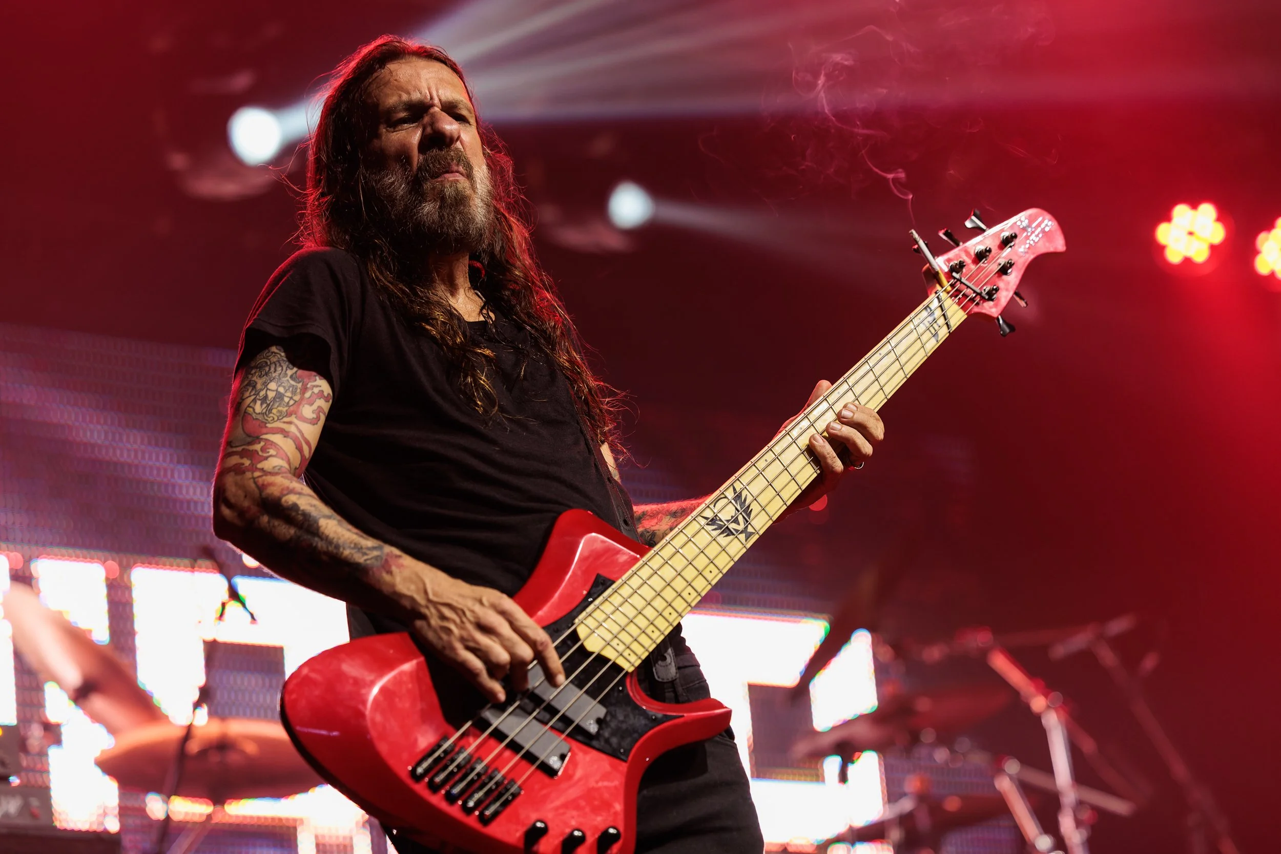 A man with long hair and a beard playing a red electric bass guitar on stage, with stage lights and a large digital screen in the background.