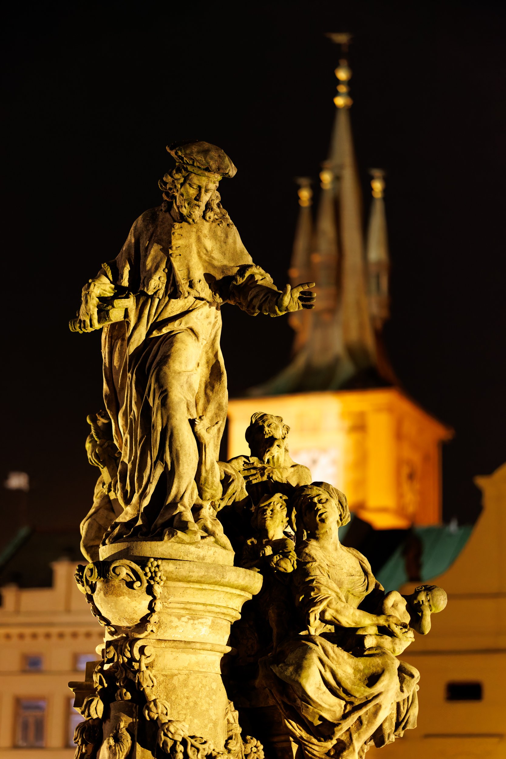 Night view of an ornate stone fountain with sculpted figures, illuminated by warm lighting, with a historic building with a steeple in the background.
