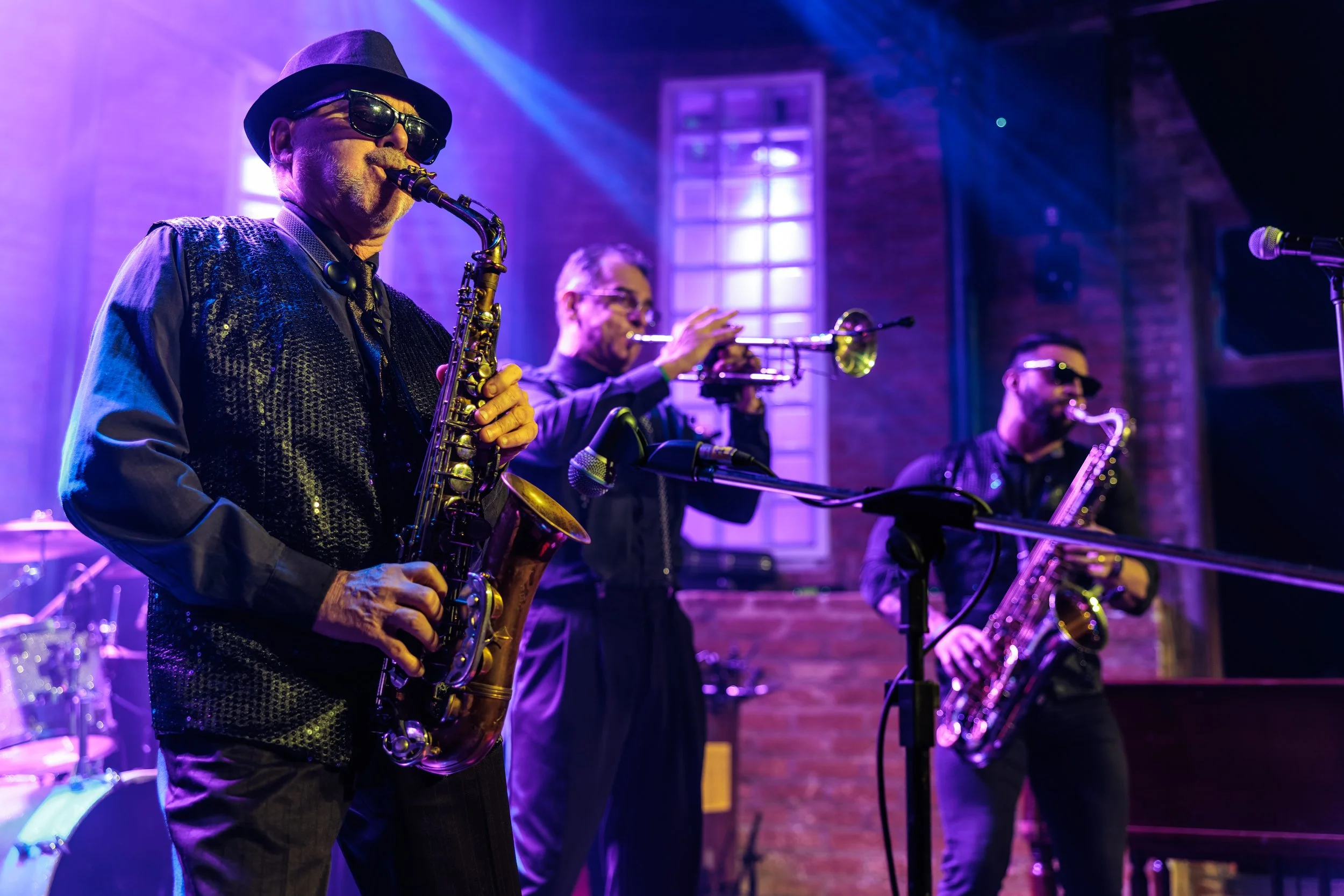 Jazz band performing on stage in a dimly lit venue with purple and blue lighting. The band includes saxophonist, trumpet player, and a bass player, all wearing dark clothing and sunglasses.