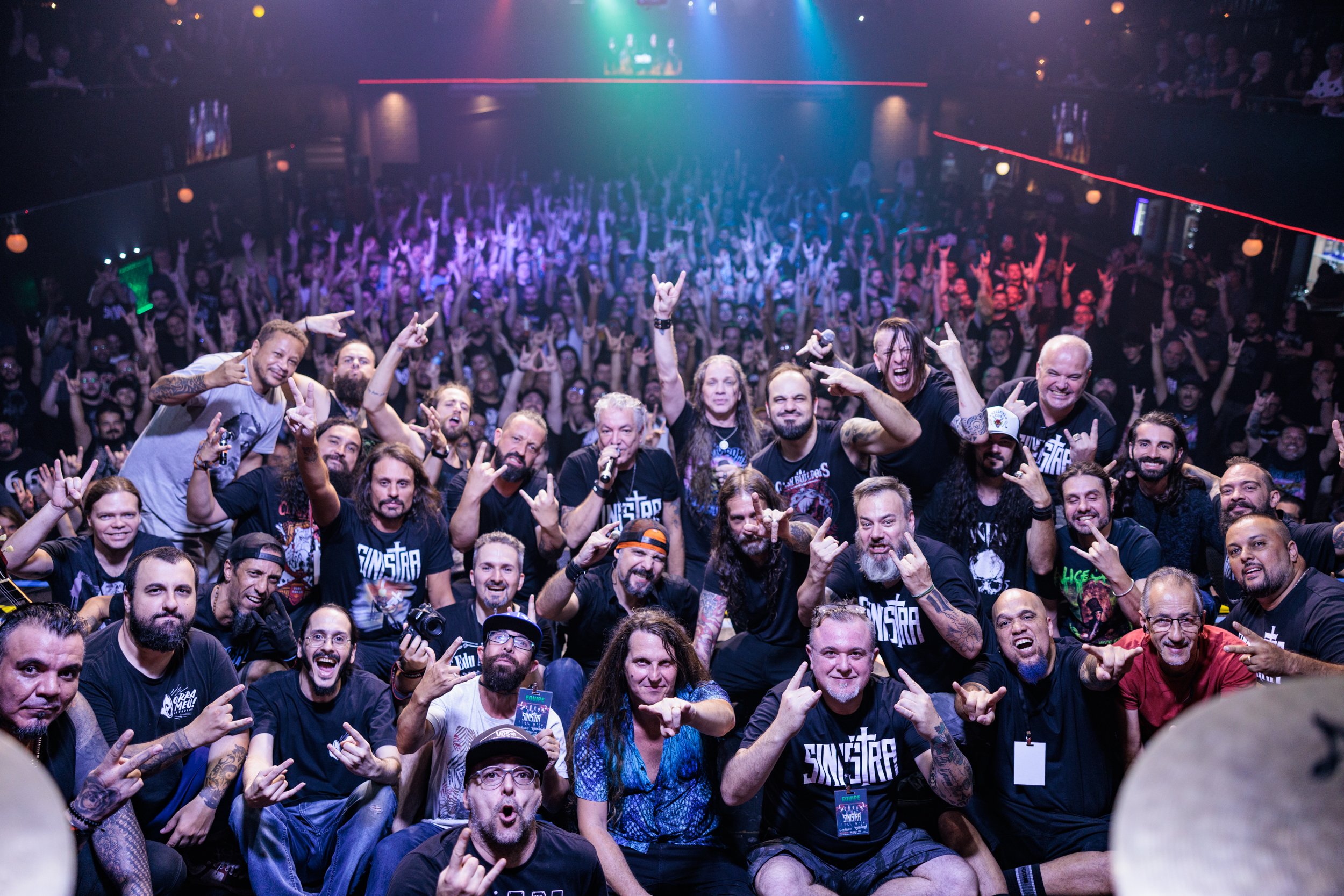 Crowd of concert attendees at a music event, many making rock hand gestures, with stage lights overhead illuminating the scene.