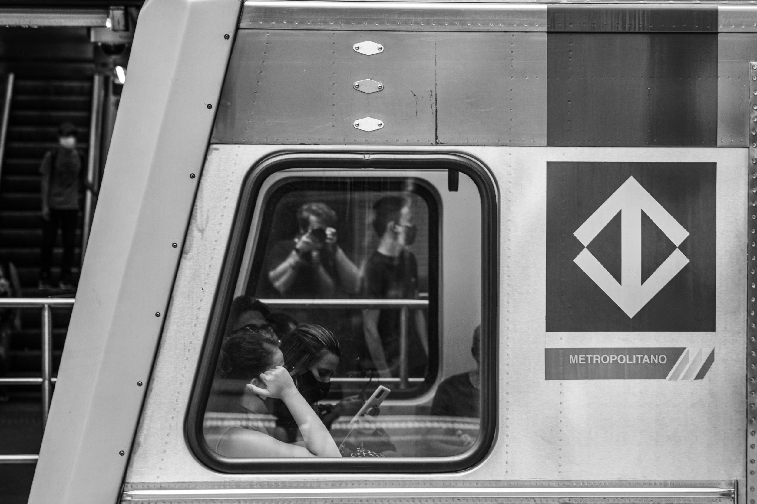 People wearing face masks sitting inside a subway train, visible through the window, with an upward pointing arrow sign on the subway exterior labeled 'METROPOLITANO.'