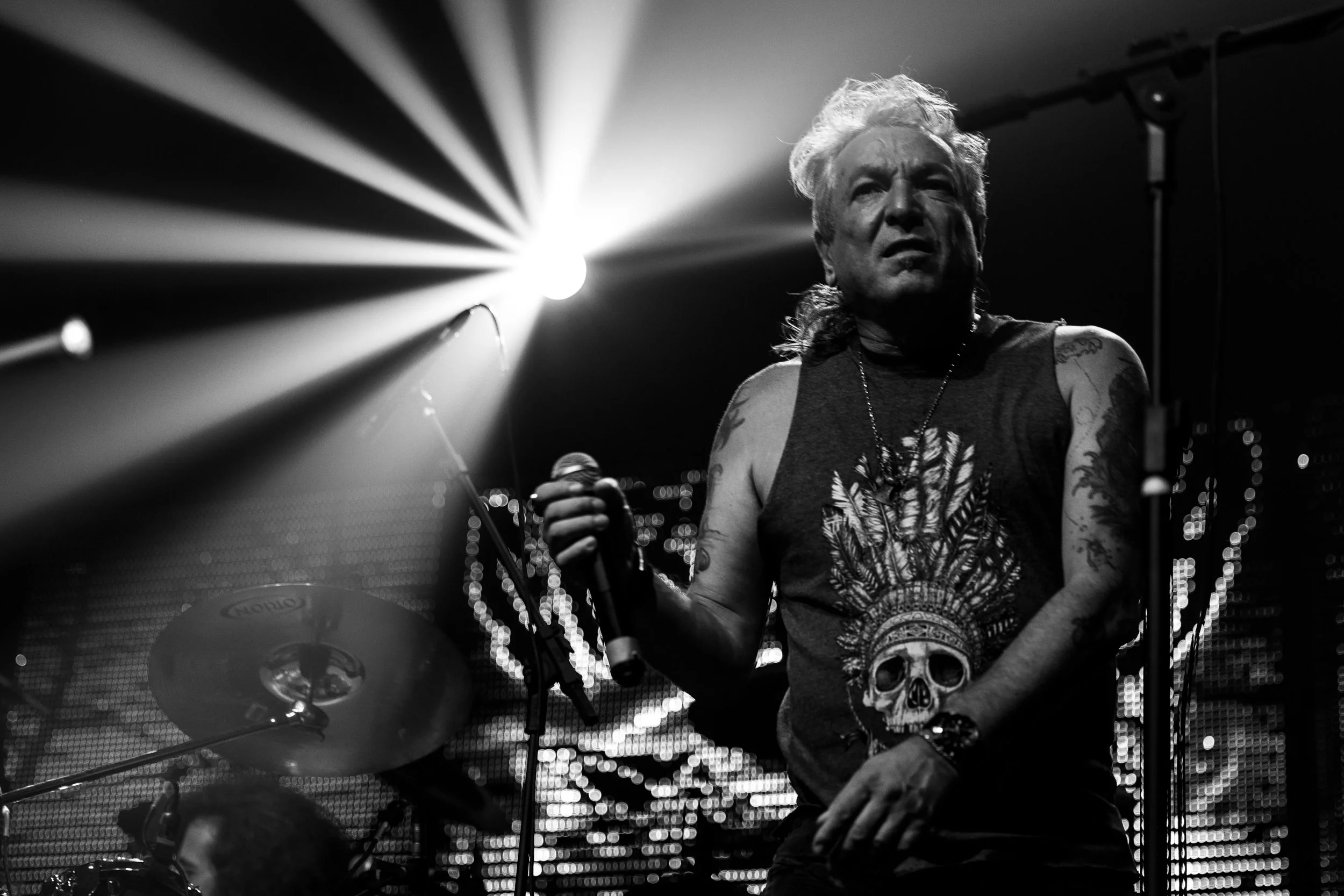 Black and white image of an older male drummer with tattoos, wearing a sleeveless shirt and holding a microphone in his right hand, performing on stage under bright lights.
