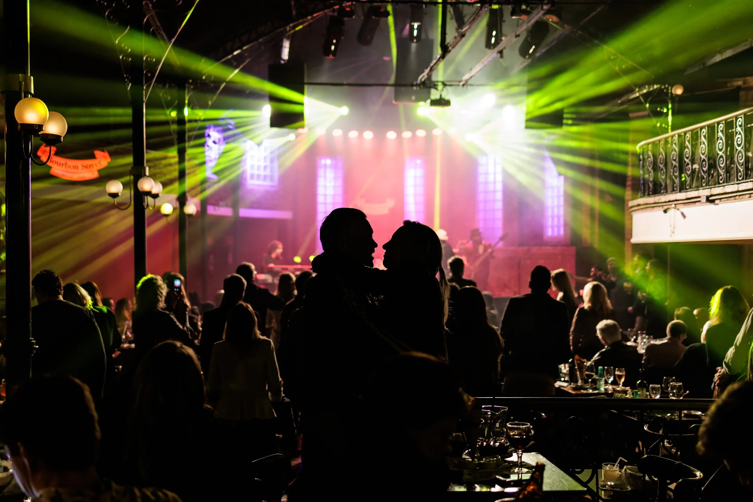 A lively indoor concert or event with green and purple stage lights shining on the crowd, silhouettes of people enjoying the show, and a stage with performers and large stained-glass windows in the background.