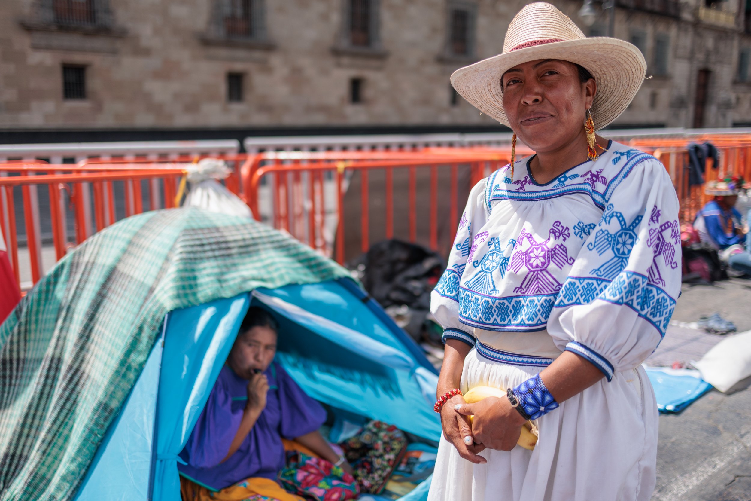 Woman dressed in traditional Mexican clothing standing next to a woman sitting inside a small tent.