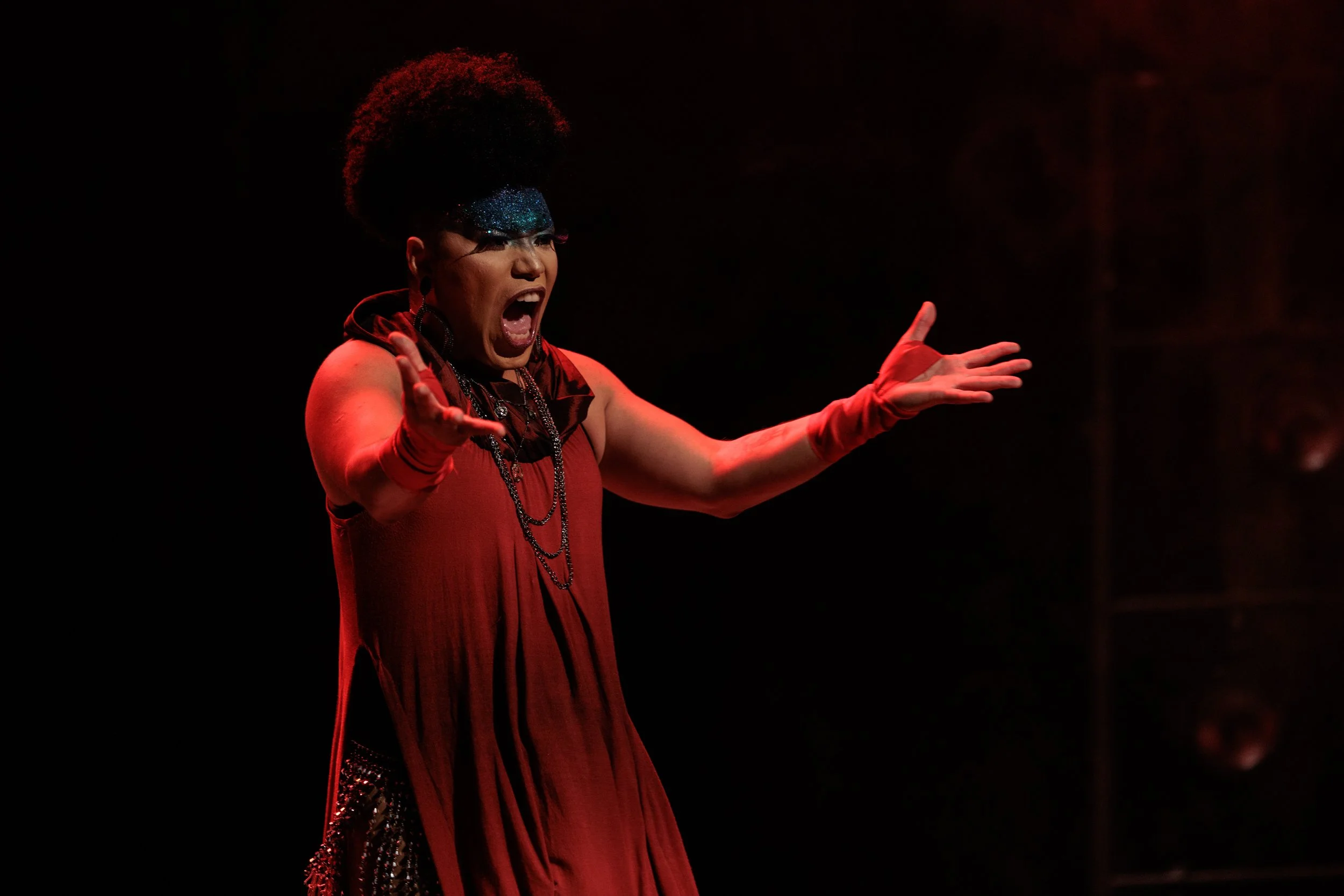A woman singing passionately on stage, wearing a red dress, long red gloves, and a sparkling blue eye mask, with curly hairstyle and expressive gesture.