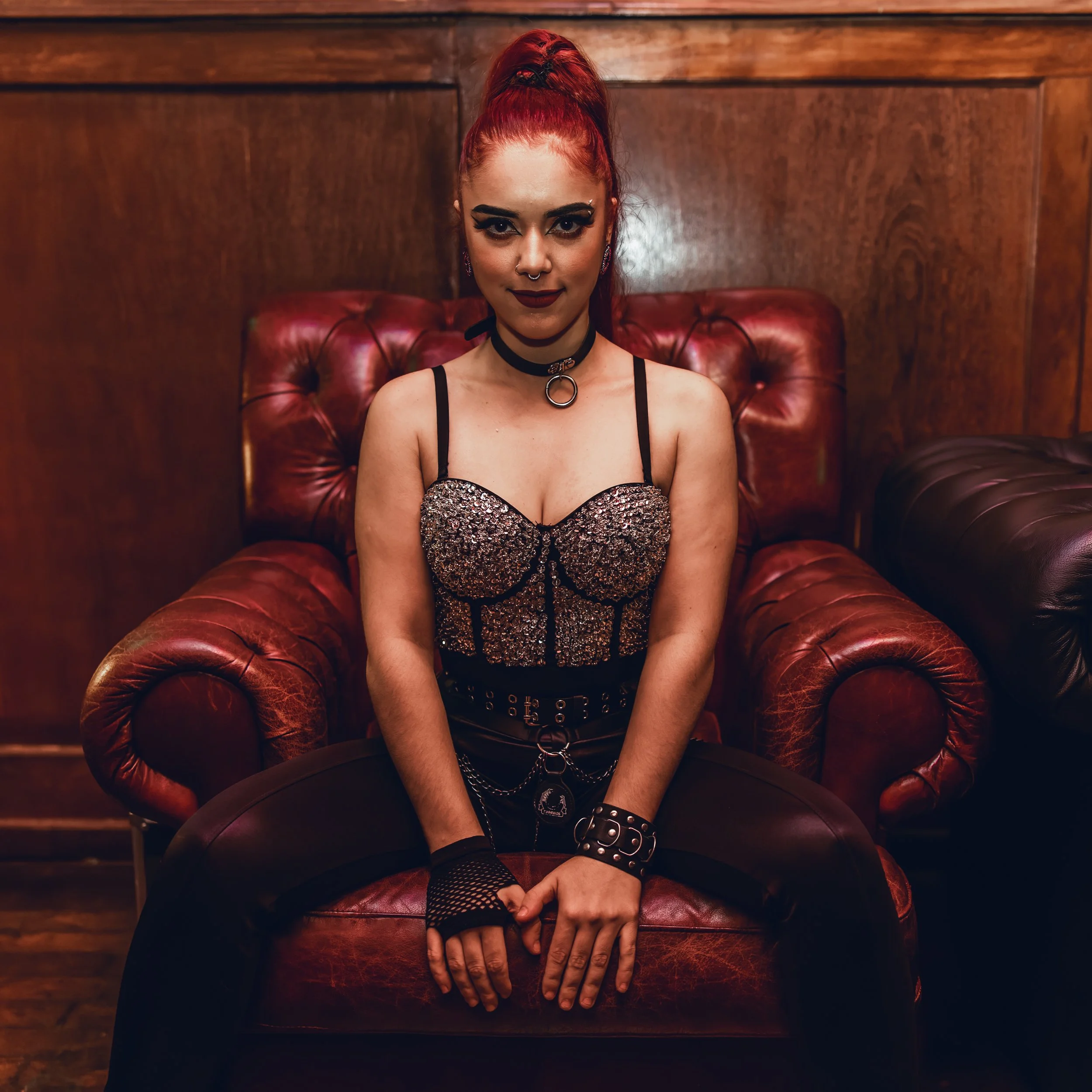 A woman with bright red hair in a high ponytail, dark eye makeup, and dark lipstick, sitting on a red leather couch with a wooden wall behind her. She is wearing a black choker with a ring, a black lace top with sparkly embellishments, and a studded 