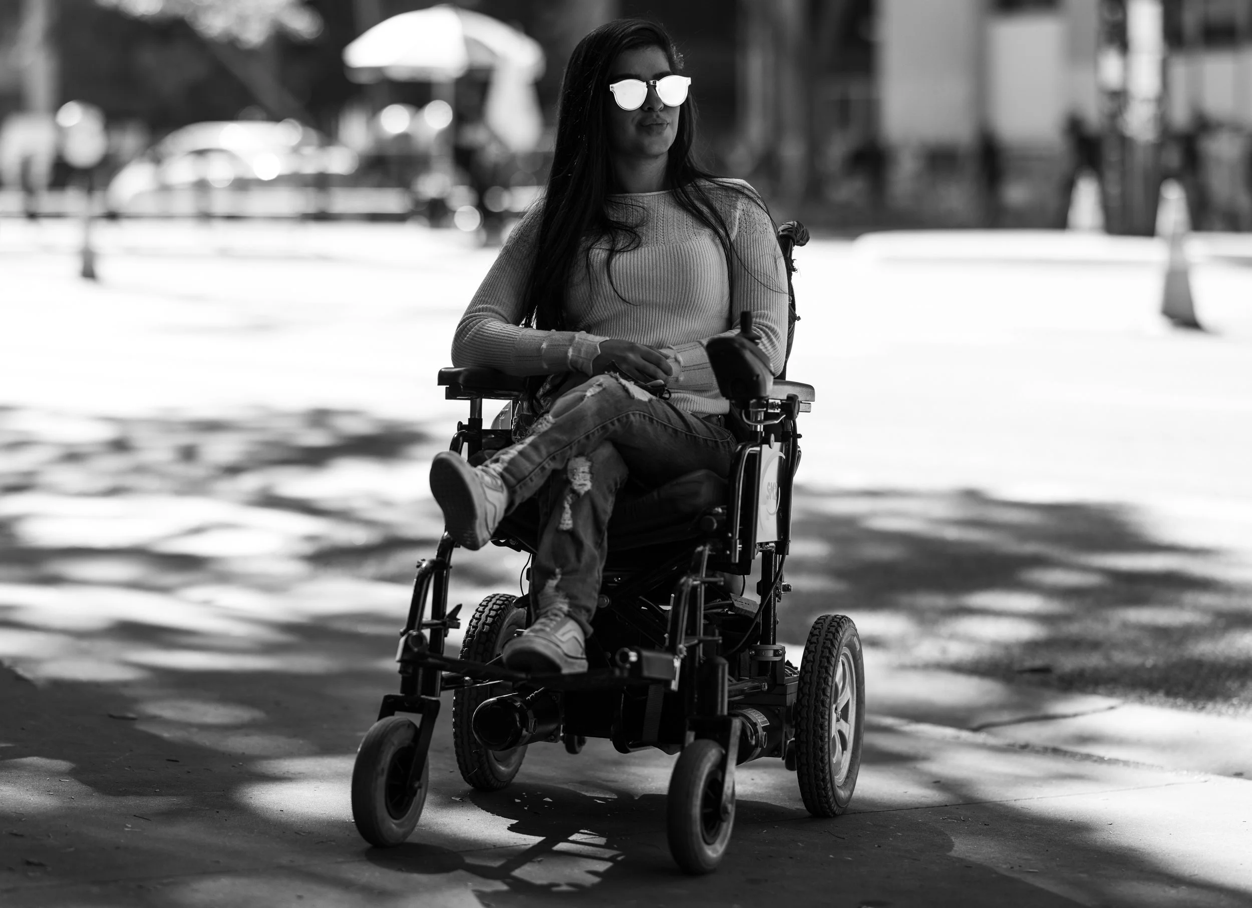A woman in sunglasses sitting in a wheelchair outdoors, with buildings and trees in the background.