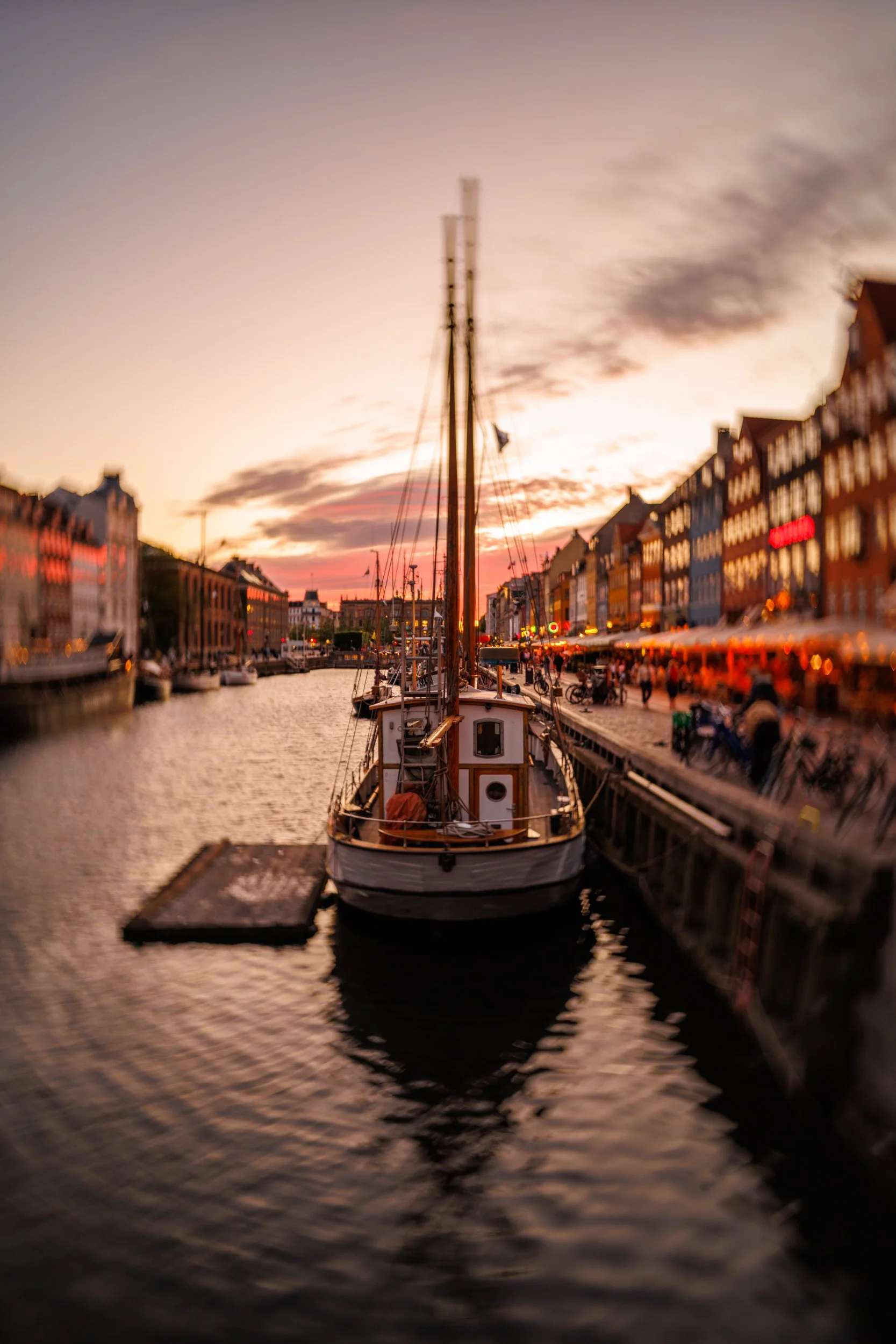 A boat docked at a harbor during sunset, with colorful sky and blurred buildings in the background.