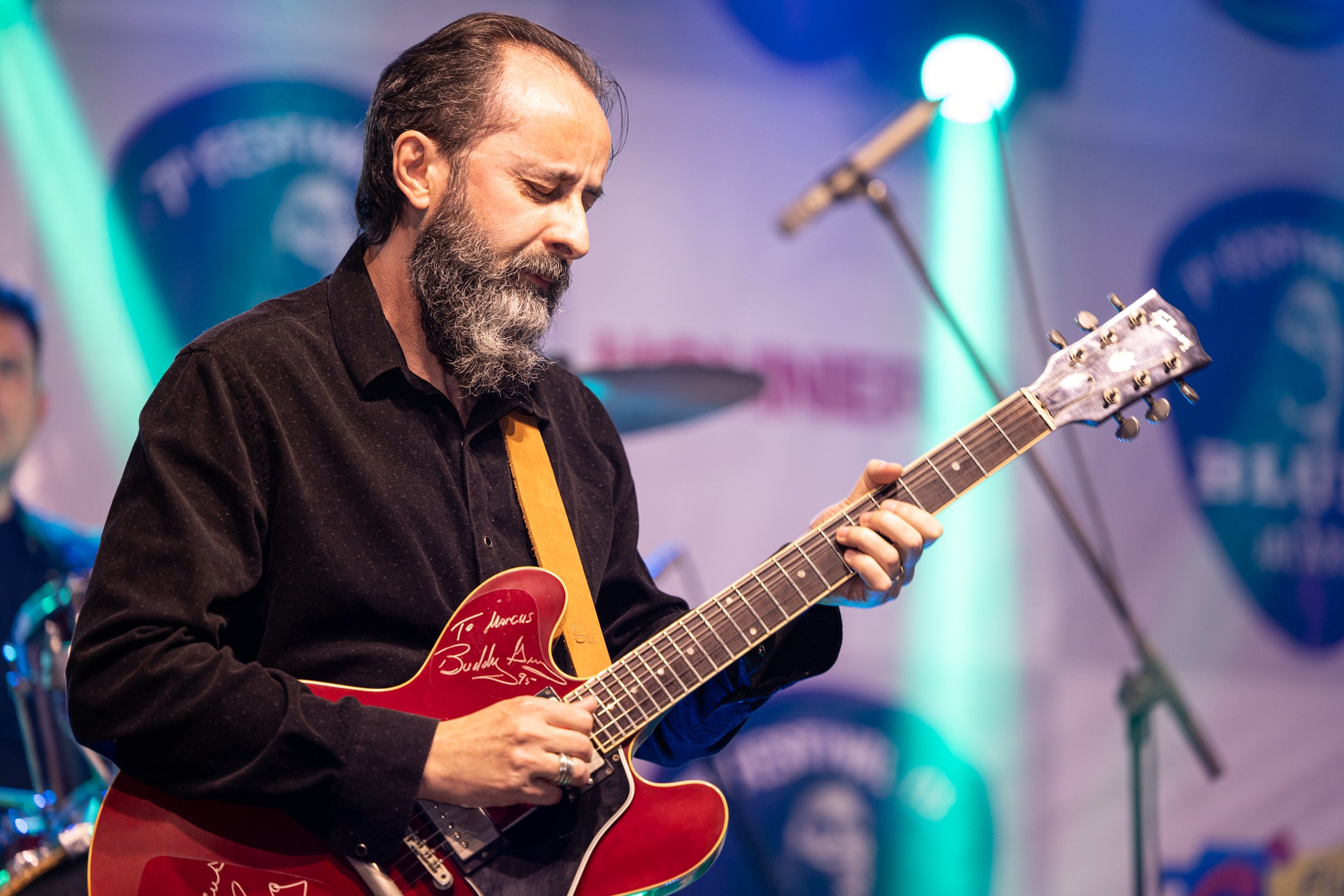 A man with a beard playing an electric guitar during a live performance, with stage lights and a blurred background.