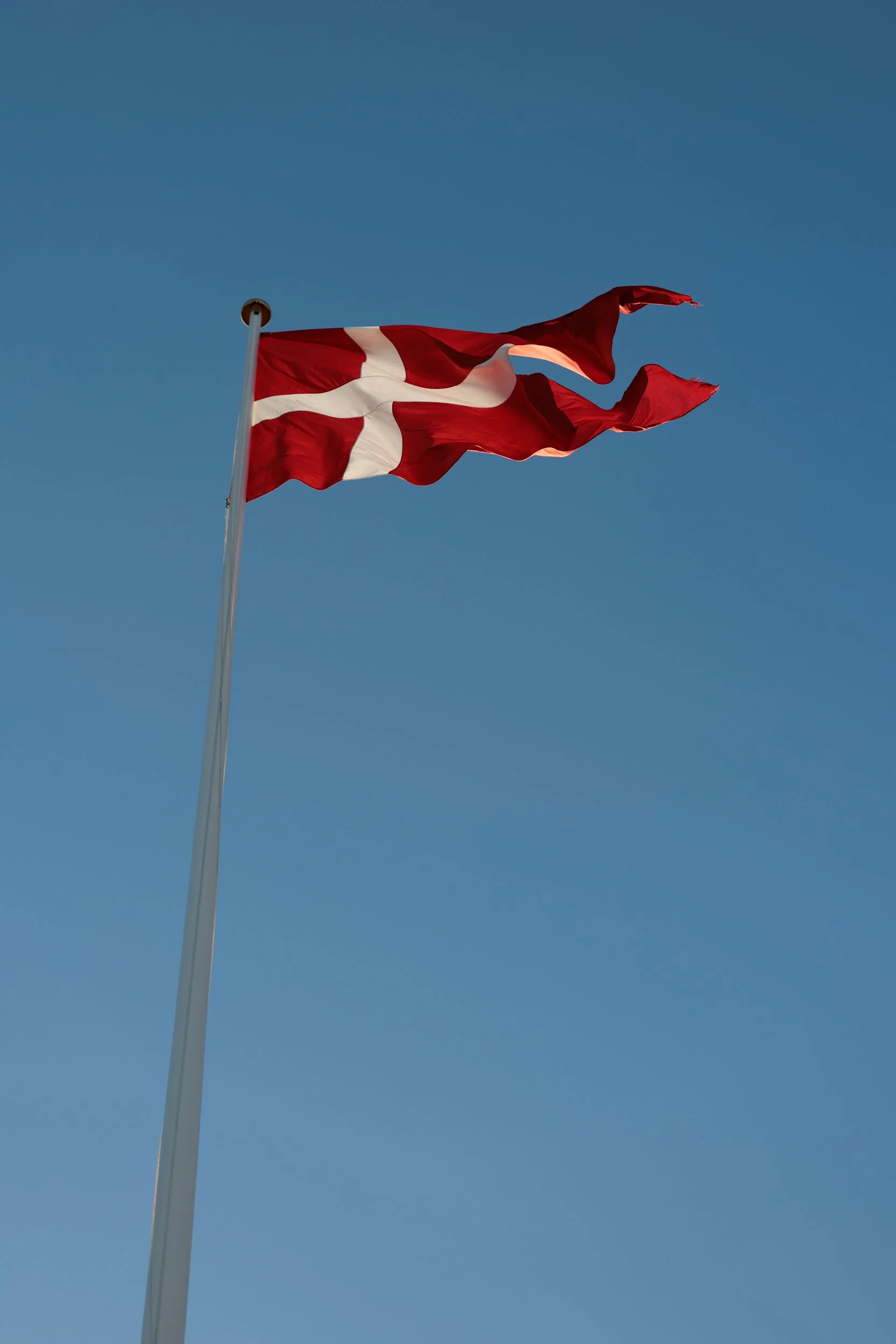 A red flag with a white Scandinavian cross flying against a clear blue sky on a tall flagpole.
