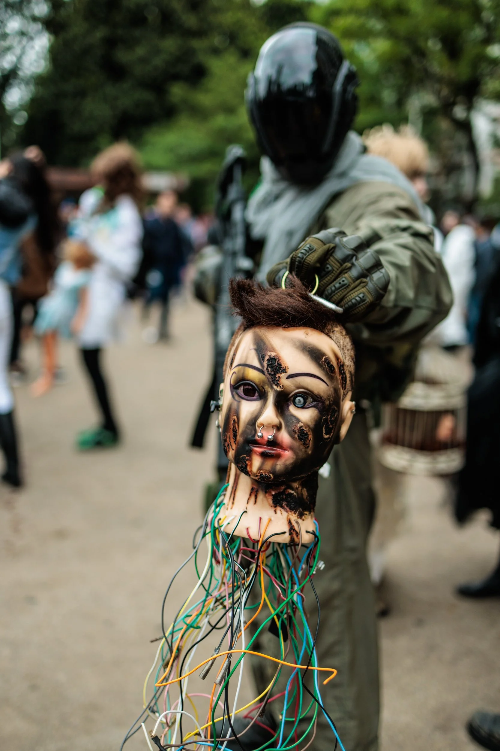A person holding a human-like mask with beads and wires hanging from the mask's face, with a blurred crowd and trees in the background.