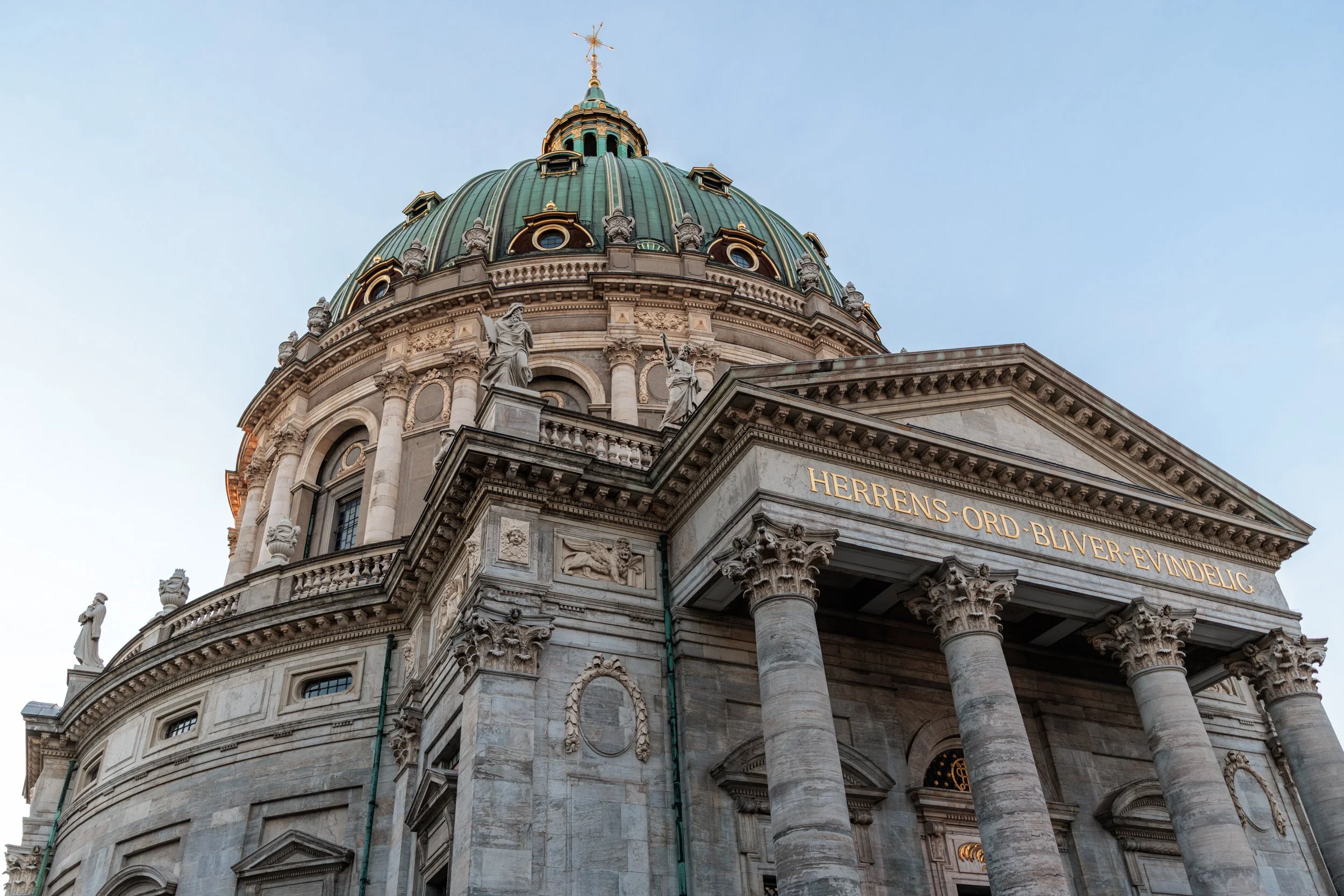 Close-up of a grand, ornate historical building with a large green dome, multiple statues, and intricate stone carvings, featuring the inscription 'HERRIENS-ORD-BIVVER-EVINDALIC' on the facade.