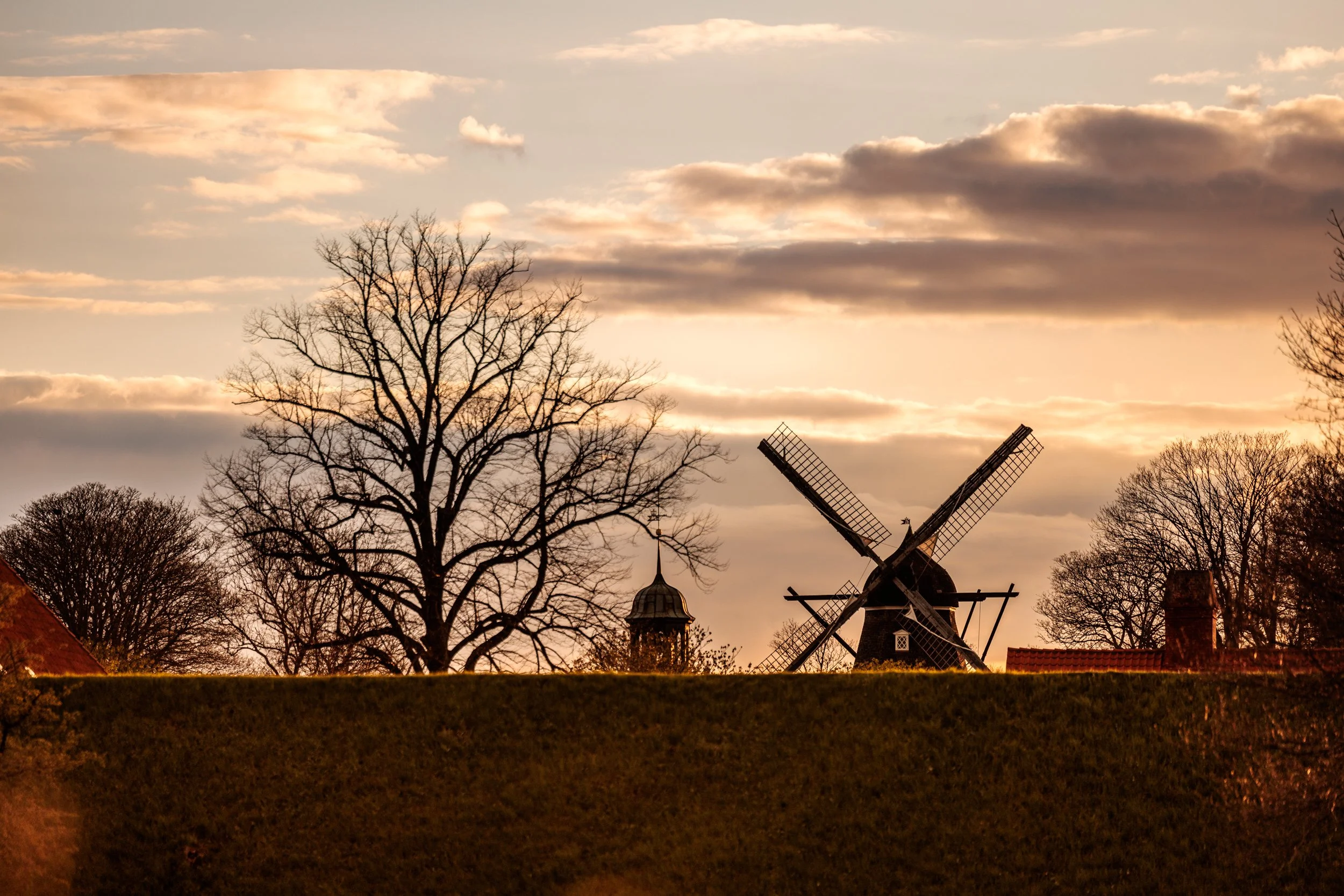 A windmill in a rural setting with leafless trees, a cloudy sky during sunset.