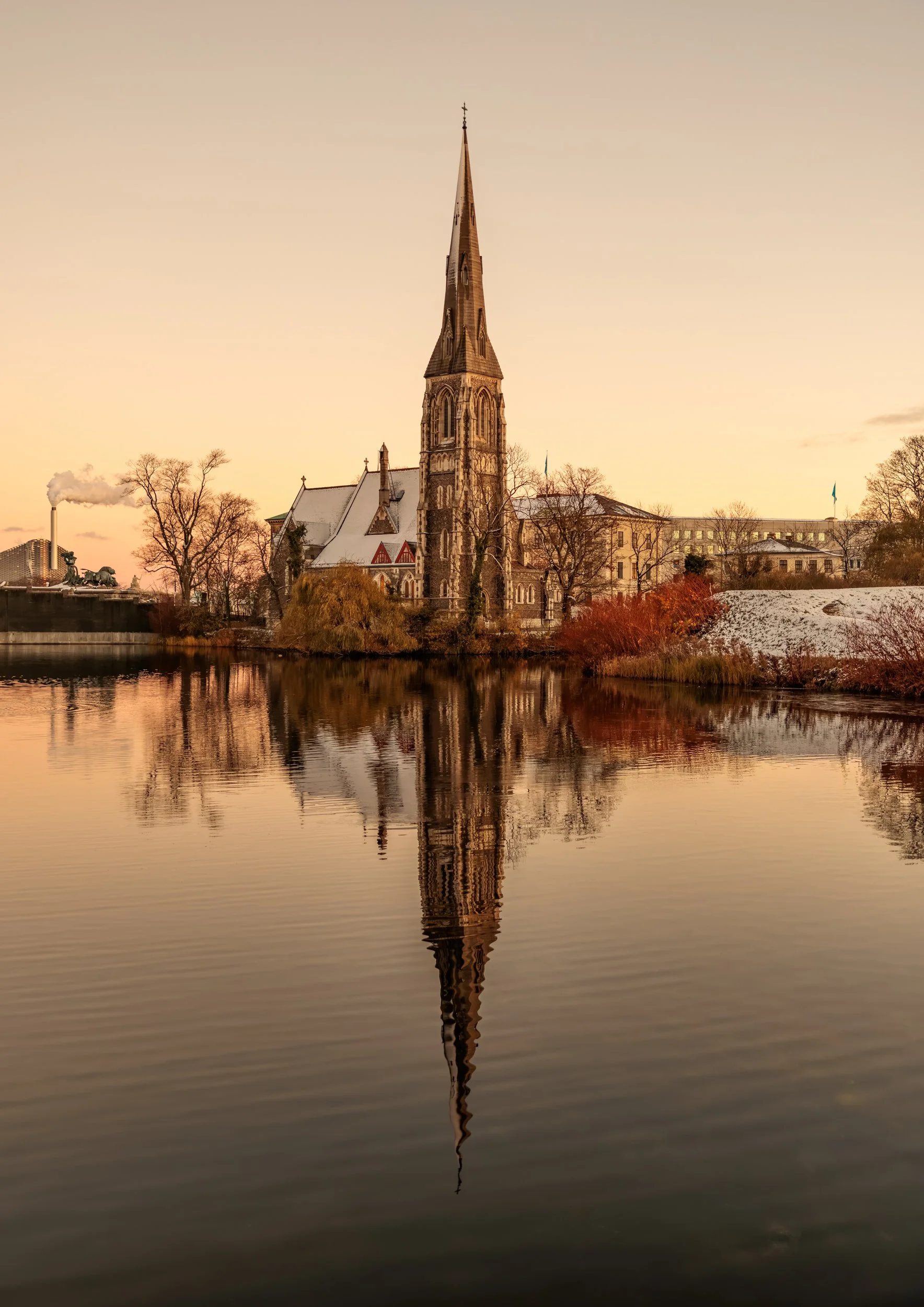 A church with a tall steeple reflected in a calm body of water, with trees and a building in the background at sunset.